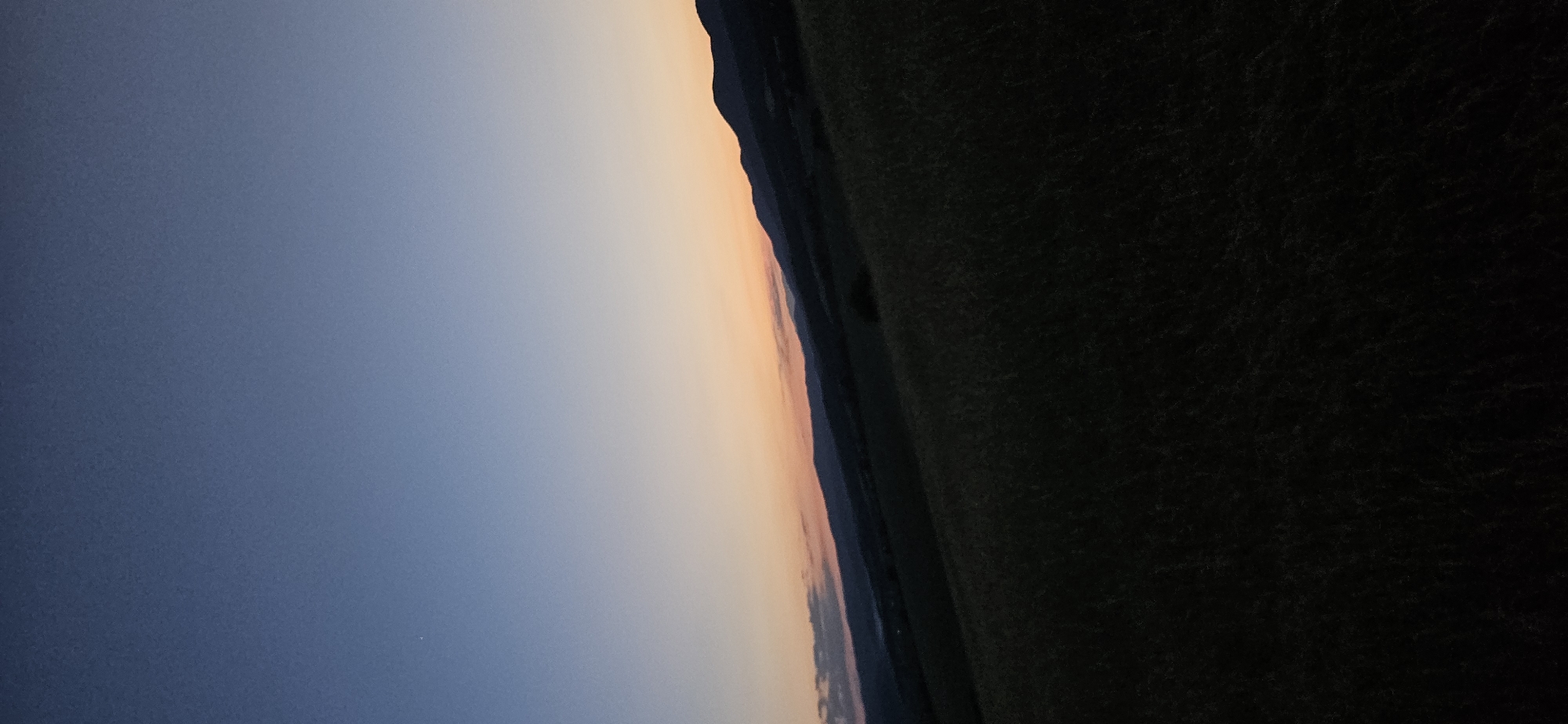 An outdoor, vertical, long shot captures a rural landscape at dusk. The sky is a gradient of deep blue at the top, transitioning to a pale orange and pink near the horizon, where a silhouetted mountain range stretches across the frame. In the foreground, a dark, grassy field fills the lower half of the image, with subtle textural details of grass blades visible. The overall scene suggests a peaceful evening in a remote, natural setting, with no people or specific structures apparent within the frame. The image does not contain any identifiable landmarks that would indicate its location in the city of Momin Prohod, Bulgaria.