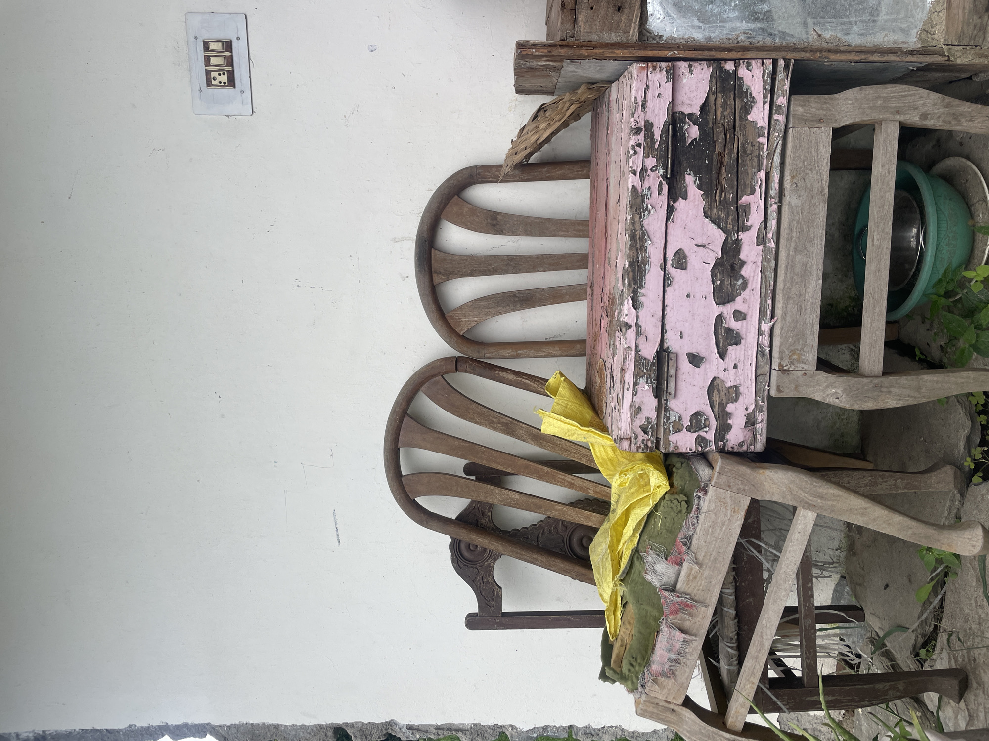 Two weathered wooden chairs are placed against a white stucco wall in Bhagsu, India. The chair on the left is older, with intricate carvings on its backrest, and is draped with a faded green cloth and a bright yellow plastic bag. The chair on the right is simpler, with a curved slatted back. Between the chairs, a distressed pink wooden chest sits atop another set of wooden legs. The chest has areas where the paint has chipped away, revealing the dark wood beneath. A dried palm frond leans against the right side of the chest. Below the chest, a green plastic basin contains a metal bowl. The wall has an electrical switch plate with three switches mounted to its right. The ground beneath the chairs is uneven and appears to be covered in debris and sparse vegetation.