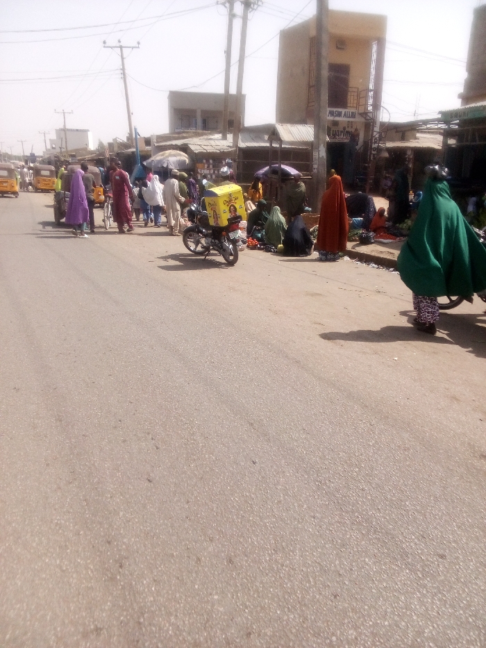 This image depicts a bustling street scene in Rangaza, Nigeria, likely functioning as a market area during daylight hours. The foreground consists of an empty, asphalt paved road with visible shadows cast from the right side, indicating direct sunlight.
In the midground, the road accommodates pedestrian traffic and parked vehicles. On the left, several yellow tricycles (commonly known as Keke Napep) are visible, along with individuals walking. One person wears a purple headscarf and long garment. Another individual walks a bicycle.
On the right side of the road, a vibrant market operates along a raised curb. Numerous individuals, predominantly women, are present. Many women are seated on the ground or low stools, displaying produce such as vegetables and fruits for sale. Their attire includes various colored head coverings and long garments, with prominent examples in green, black, and rust/orange. One woman, clad in a green full-length covering over patterned pants, walks away from the viewer on the far right.
A black motorcycle equipped with a large, yellow rectangular delivery box featuring "ChapChap" branding is parked on its kickstand on the right side of the road, amidst the market activity.
The background features a collection of buildings, mostly one or two stories high, constructed from light-colored materials. One notable two-story building on the right has "MASIN ALLAH" and "iyarin" painted on its facade. Multiple utility poles with a dense network of overhead wires line the street, extending into the distant background. Several makeshift stalls with corrugated metal roofs and some white market umbrellas provide shade for vendors. The overall environment suggests a lively commercial district under clear, sunny skies.