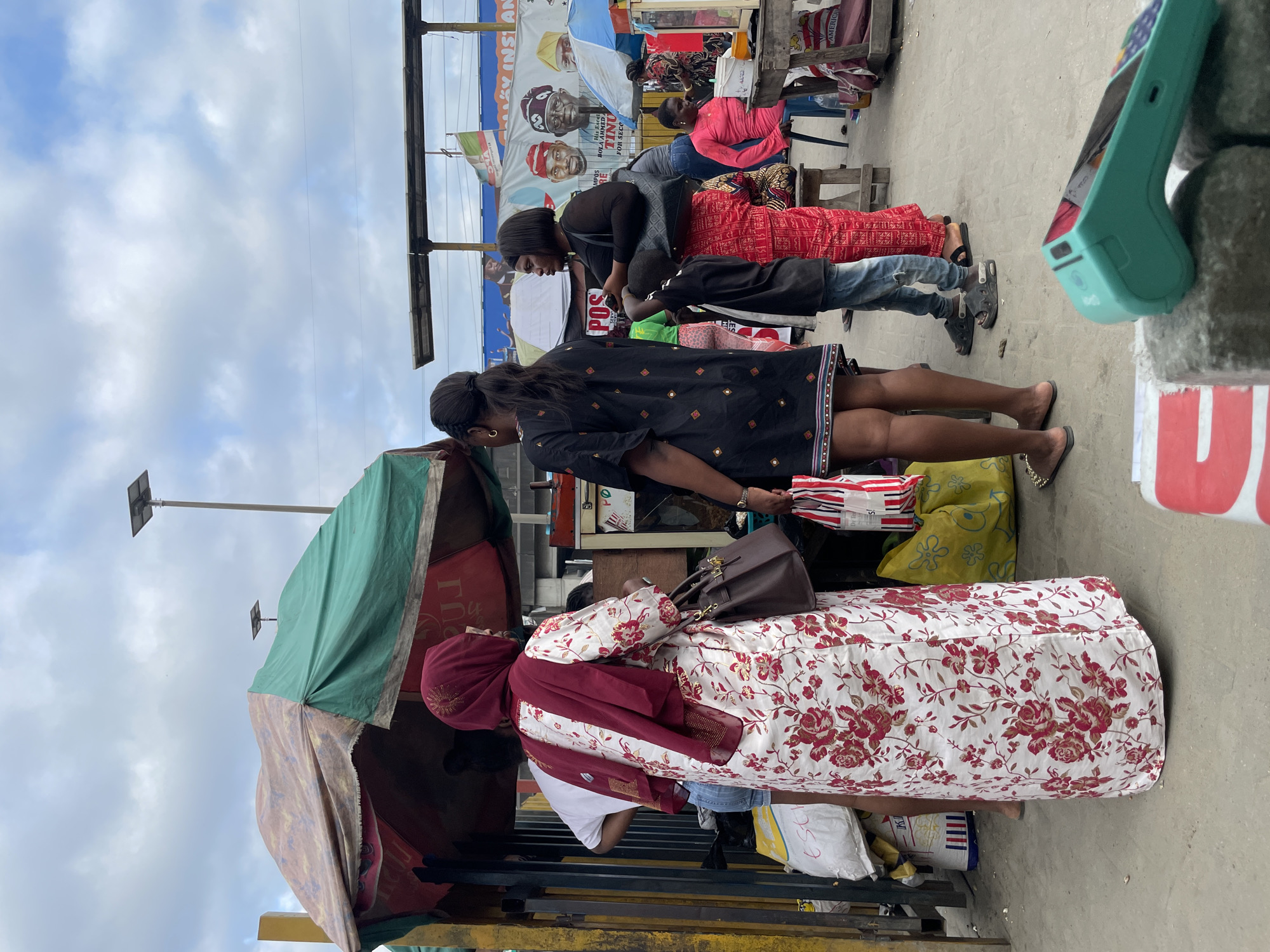 In Lekki, Nigeria, a street scene unfolds under a cloudy sky. A woman wearing a red hijab and a white dress with red floral patterns stands facing away from the camera, her back towards a market stall covered by a striped and green tarp. She carries a brown handbag. To her left, another woman in a black dress with geometric patterns stands with her back to the camera, holding a red and white striped bag. A young boy in a black t-shirt and blue jeans stands to the right of the woman in the black dress, facing left. Further to the right, a woman in a black top and red patterned pants looks towards the left of the frame. Behind her, a woman in a pink long-sleeved shirt sits at a table. Various market goods and stalls are visible in the background, including a large yellow bag and a white sack. A blue umbrella is positioned behind the woman in the pink shirt. A promotional banner with images of men is displayed in the distance. A teal-colored point-of-sale device lies on a rough surface in the foreground.