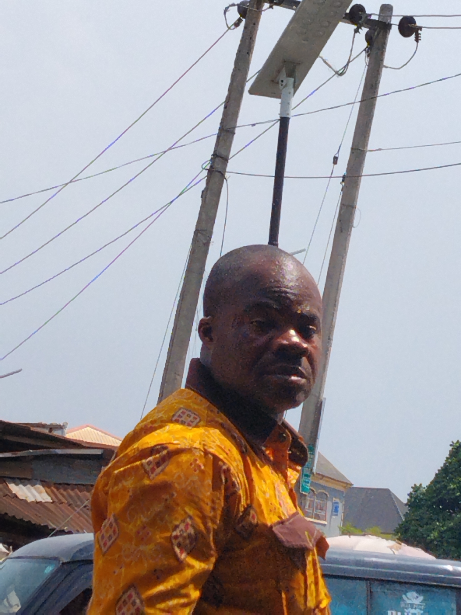 A medium shot shows a dark-skinned man with a shaved head wearing a patterned yellow shirt. The man is centrally located and slightly angled to the left. He is looking towards the viewer with a serious expression. Behind the man are two utility poles with electrical wires running between them. One of the poles has a modern LED street light attached. In the background, cars are parked and houses are visible. A corrugated metal roof is seen above a car. Based on the architecture and the presence of utility poles and cars, the location is likely an urban setting. The location is specifically within the city of Aba, Nigeria.