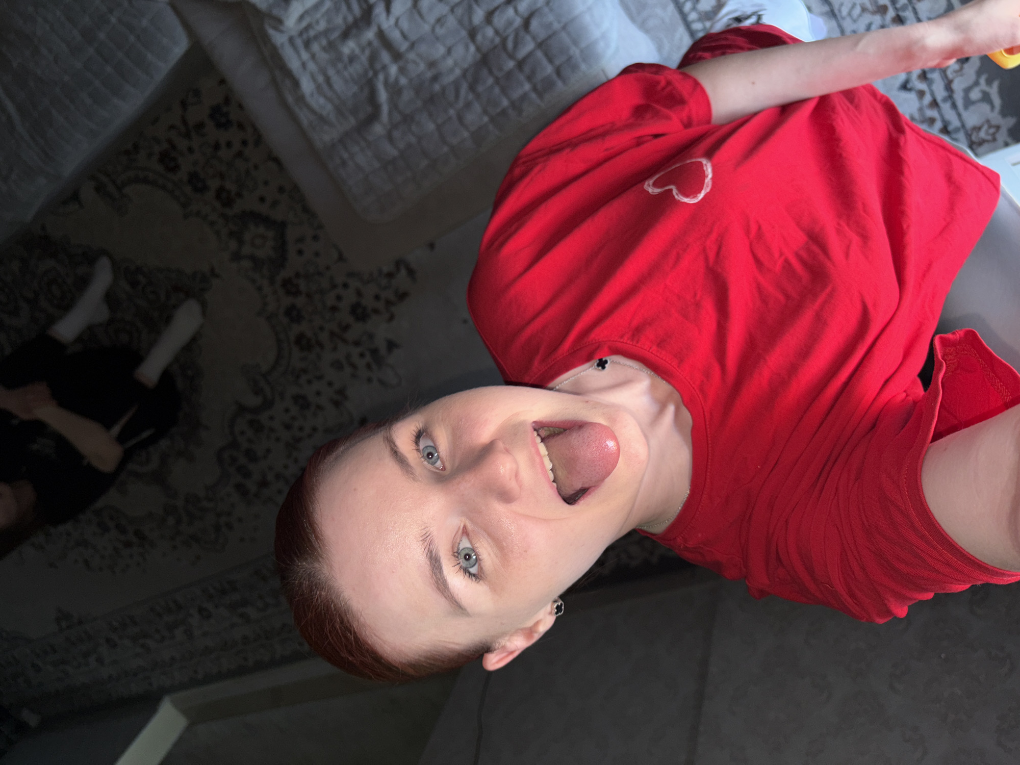 A selfie taken from a low angle shows a young Caucasian woman with red hair pulled back. She is wearing a bright red t-shirt with a white heart design on the left side of the chest. Her mouth is open and her tongue is extended. Her eyes are open and looking at the camera. She is wearing small black earrings. In the background, a patterned rug is visible, and further back, a bed with white quilted bedding. Another person, wearing black clothing and white socks, is visible in the background, sitting on the rug. The scene appears to be indoors, possibly a bedroom or living area. The lighting is dim, suggesting it may be nighttime or a dimly lit room.