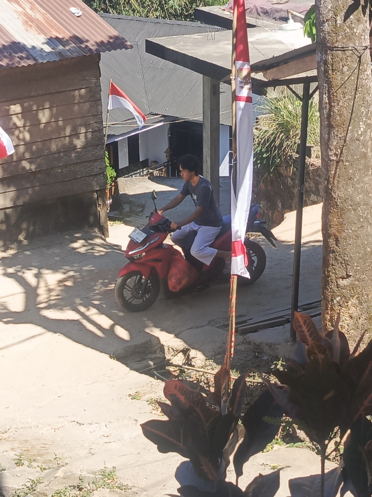 A young Indonesian man is riding a red scooter on a dirt path. He is wearing a dark t-shirt and white pants. The scooter has a license plate that reads "90 5419 GI". There are two Indonesian flags visible in the scene, one is draped over a bamboo pole in the foreground, and another is flying from a pole in the background. In the background, there are several wooden buildings and a glimpse of lush green vegetation. The ground is uneven and dusty, with shadows cast by sunlight filtering through trees. The overall scene appears to be in a rural or semi-rural setting in Kendari, Indonesia.