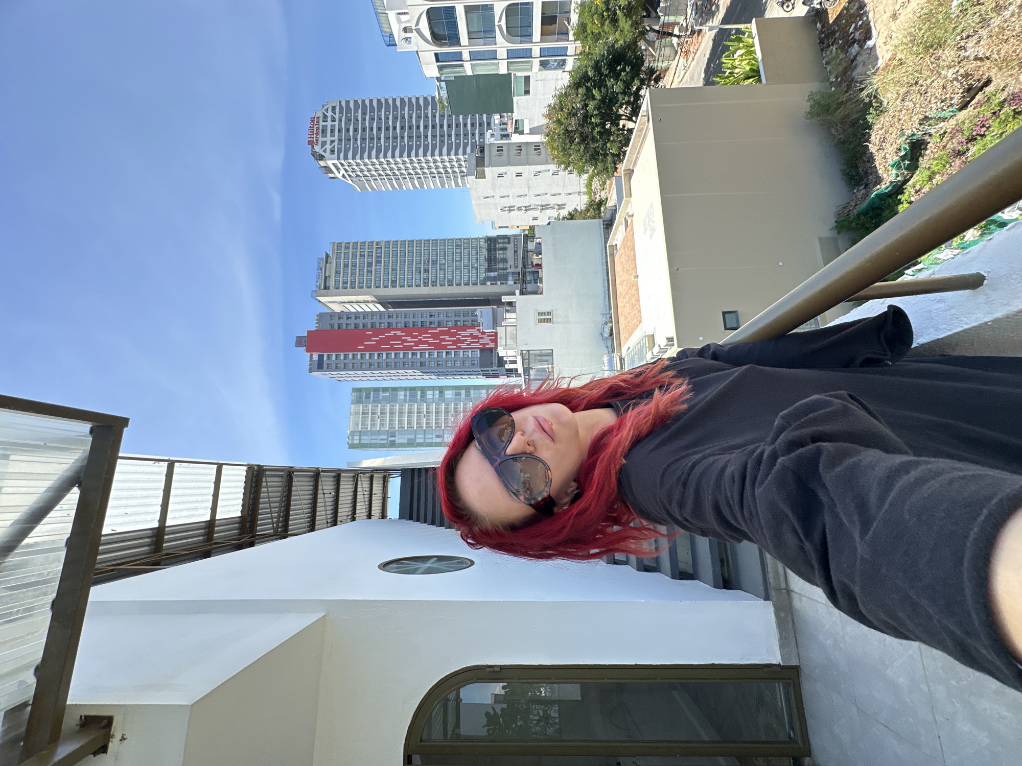 A woman with vibrant red hair and large sunglasses is taking a selfie on a balcony overlooking a city. She is wearing a black long-sleeved shirt. In the background, several tall buildings are visible, including the Hilton Garden Inn and buildings with red and white facades. The sky is clear and blue. The balcony has a railing and a corrugated metal roof overhead, supported by metal beams. Adjacent to the balcony is a white building with a dark arched doorway and a circular window. The scene appears to be in the city of Đà Nẵng, Vietnam.