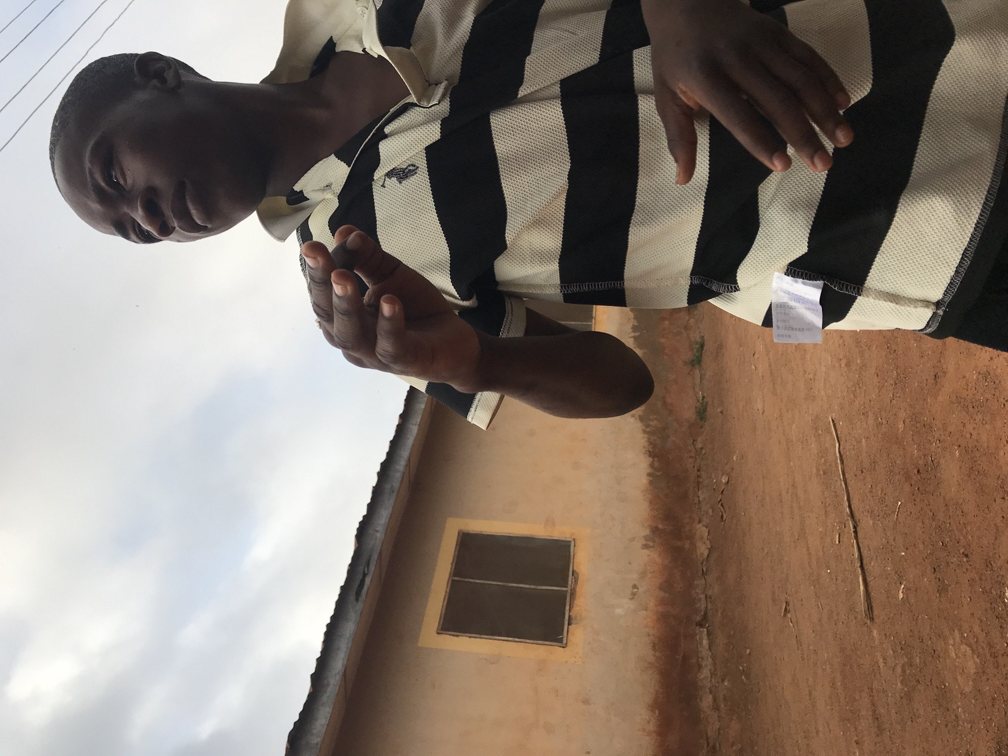 The image displays a young person with dark skin and short, dark hair, standing outdoors during daytime. The individual is wearing a black and white striped polo shirt and is looking downwards and slightly to their left with a neutral expression, holding their hands loosely in front.
In the background, to the person's right, there is a building with an earth-toned, possibly weathered, wall. A simple, dark-framed window with a grid pattern is visible on the building's facade. Above the building, a light, overcast sky suggests a cloudy day. The ground in the lower right portion of the image is reddish-brown earth, with a thin stick lying upon it. This outdoor setting, characterized by the earthy ground and the building style, is consistent with a location such as Ilorin, Nigeria.
Notable details include a small, embroidered emblem, resembling a polo player logo, on the left chest of the person's shirt. A white garment tag with illegible text, appearing to be in an Asian script with some numbers, hangs from the bottom hem of the shirt. The scene captures a casual, everyday moment in an outdoor environment.