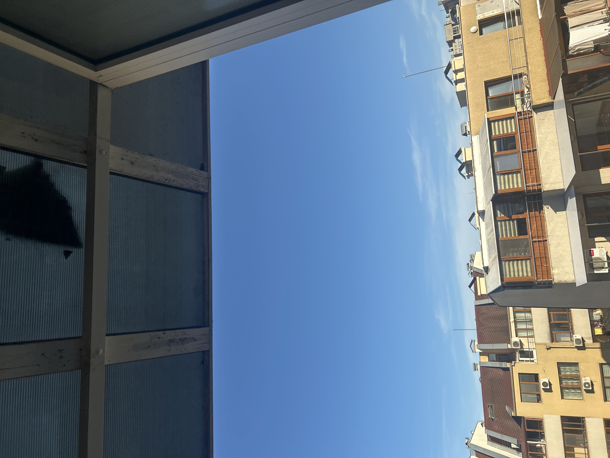 The image captures a view from a building's balcony in Varna, Bulgaria, looking out towards the sky and neighboring structures. In the foreground, a portion of a building's roof or awning is visible, constructed from corrugated plastic panels supported by wooden beams. The sky is a clear, vibrant blue, with a few faint white clouds streaking across it.
Below the sky, several apartment buildings are depicted. The buildings exhibit a mix of architectural styles and colors, with yellow and cream facades being prominent. Many have multi-story balconies, some enclosed with wooden framing and blinds, while others are open with metal railings. Air conditioning units are visible on the exteriors of some buildings. Rooftops are varied, with some covered in dark shingles and others featuring multiple chimneys or ventilation pipes. The scene suggests a daytime view in an urban environment.