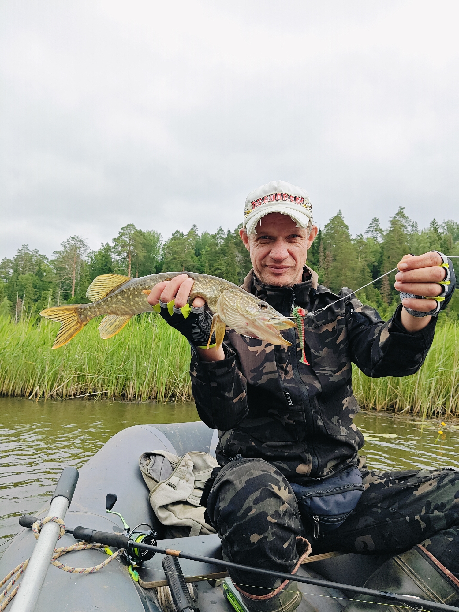 A Caucasian man in camouflage clothing and a white baseball cap sits in an inflatable boat on a body of water. He holds a freshly caught pike in his right hand and a fishing lure with multiple hooks in his left. A fishing rod and reel are visible in the boat, along with a backpack. The background consists of a dense forest and tall grass. The sky is overcast. The image appears to be taken outdoors in a rural or natural setting.
