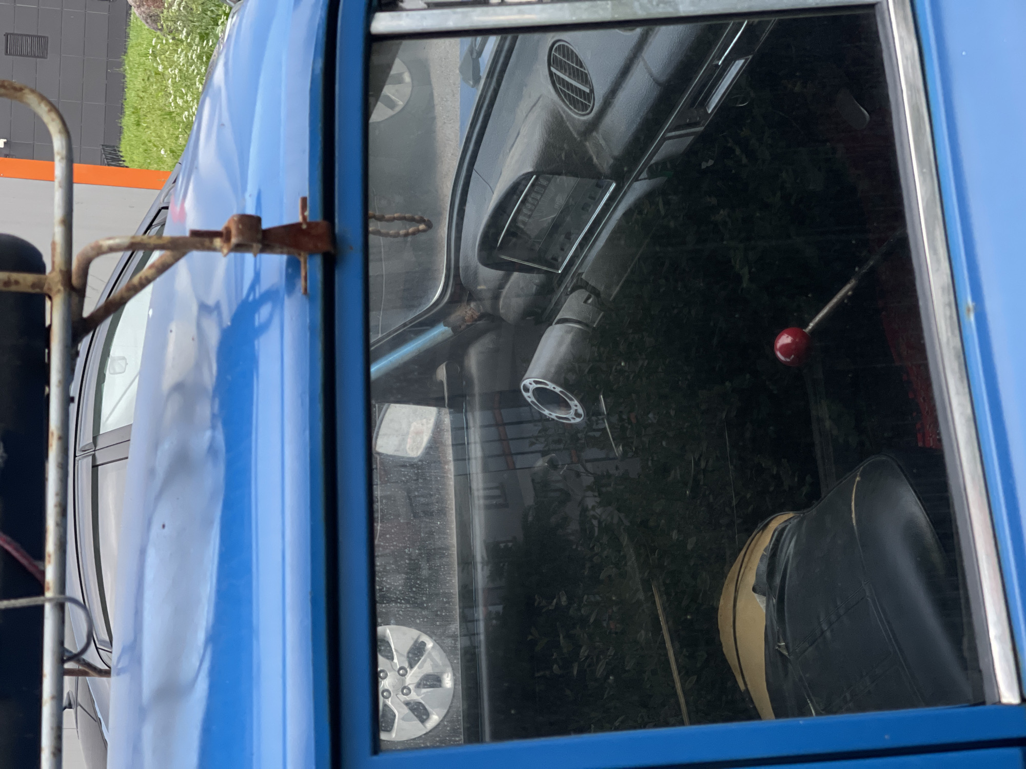 The image shows the side of a blue car, seen from the exterior. A rusty roof rack is attached to the roof of the car, with a beaded string hanging inside the car from the interior. The car has a black interior, with a steering wheel, dashboard, and a gear shift with a red knob visible through the window. The car is parked in an outdoor location, possibly a parking lot, with a building and some greenery in the background. The car appears to be old and possibly in disrepair.