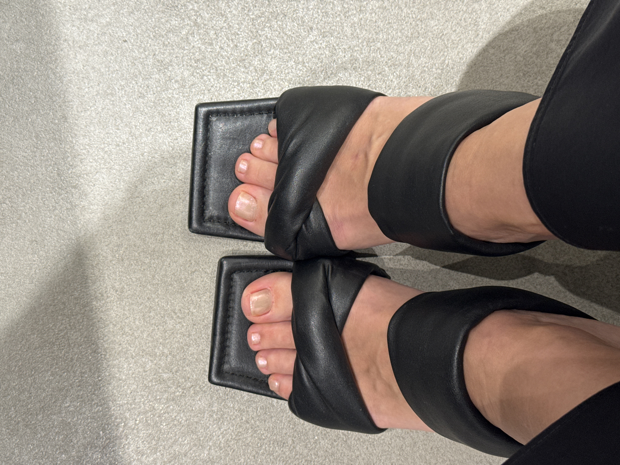 A top-down, medium-shot photograph depicts a pair of feet wearing black leather square-toe sandals. The sandals feature a wide, criss-cross strap over the top of the foot. The feet are fair-skinned, with slightly pale toenails. The subject is also wearing black pants. The background is a uniform, light gray surface, possibly tile or concrete. The setting does not appear to be in Dubai.