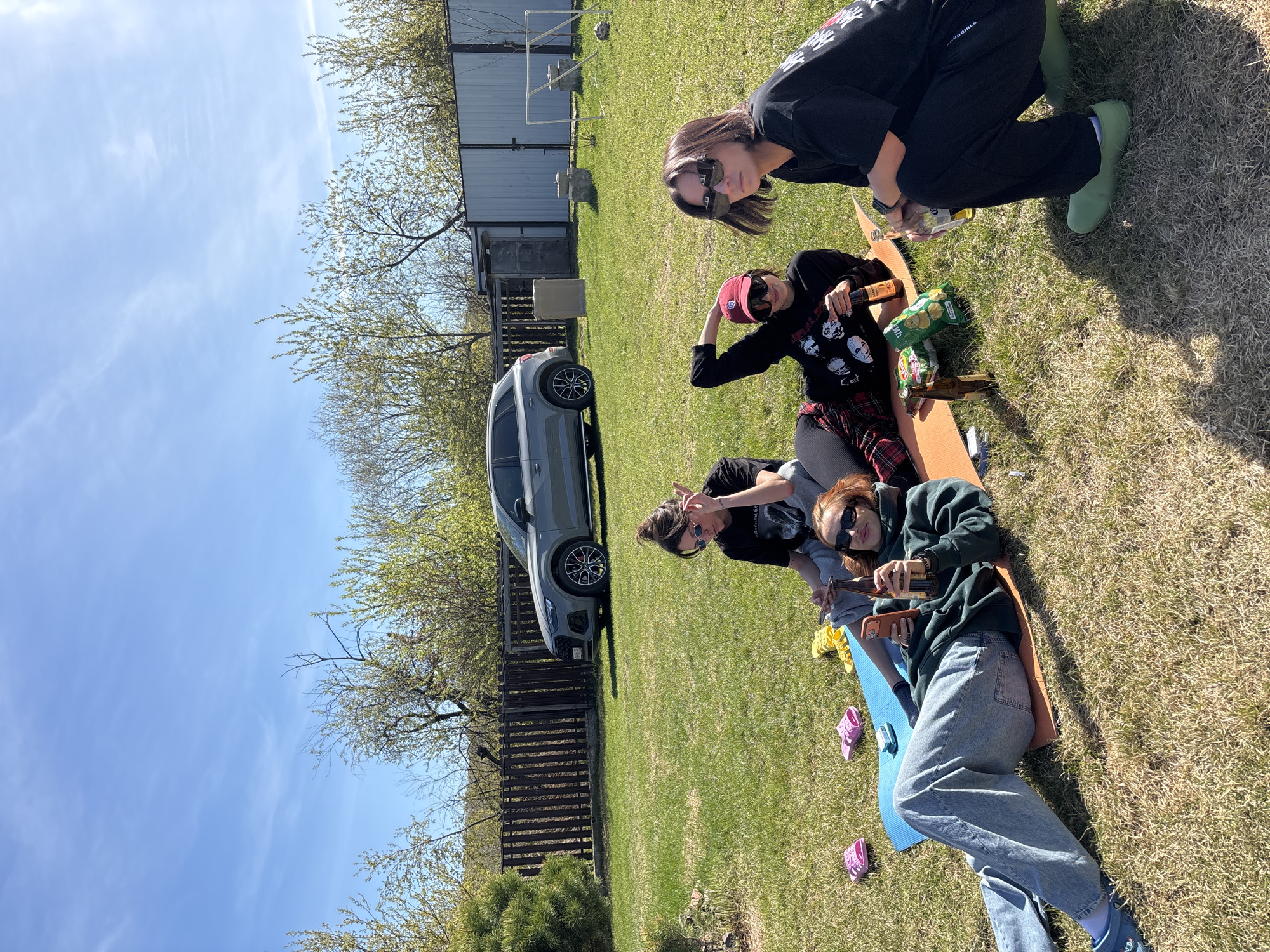 The image shows a group of five young adults gathered on a grassy lawn next to a grey car. A wooden fence and trees line the background, with a partially visible building in the distance. The sky is blue with scattered clouds.
The individuals are positioned on what appears to be a makeshift raft or sled, constructed from boards. They are casually dressed, with some wearing sunglasses and hats. Some are holding alcoholic beverages, specifically beer bottles. There is a bag of potato chips present.
The photo was taken during the daytime, and the scene appears to be a relaxed, social gathering. The precise location in Emel'yanovo cannot be determined from the image.