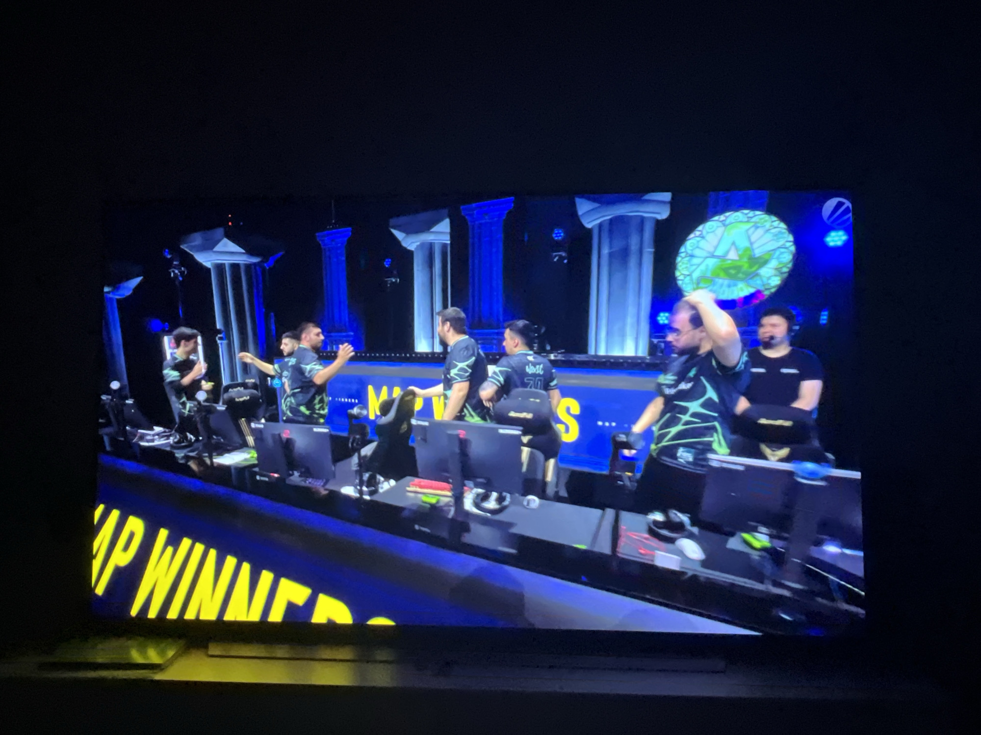 The image shows a group of young men on a stage, likely at an esports event. They are seated at a row of computers, each with a monitor. The backdrop features large, stylized white columns illuminated with blue light, suggesting a grand or ceremonial setting. Some of the men are wearing dark t-shirts with a distinctive green, geometric design. One man on the right is holding a circular, green object with a lighter green design on it, possibly a trophy or prop. Another man to his left is wearing headphones. In the foreground, a large banner with yellow text on a blue background is visible. Part of the text reads "AP WINNE". The lighting is dramatic, with spotlights and the blue illumination from the columns creating a strong contrast with the dark background and the subjects. The overall impression is of a competitive gaming event, possibly a championship, given the banner text. The image appears to be taken from a viewing angle, possibly from a spectator's seat. The date or time of day is not discernible. There are no obvious weather cues, and the location context of Varna, Bulgaria, cannot be directly inferred from the visual elements within the frame itself. The expressions on the faces of the men are not clearly visible due to the distance and image quality, but the context suggests excitement and focus.