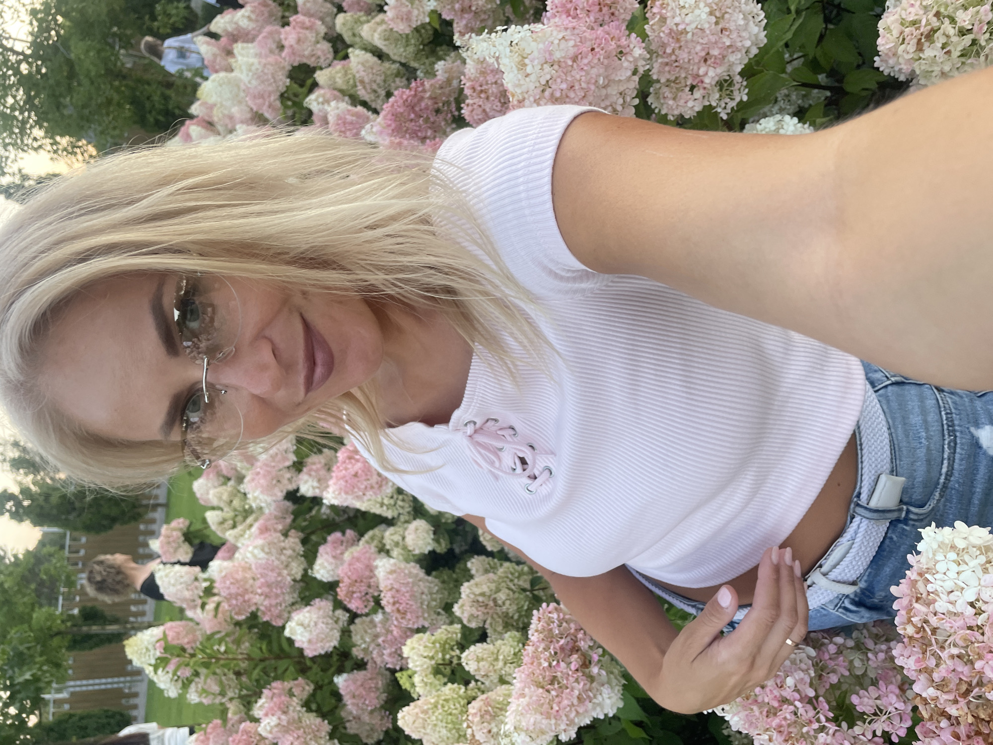 An outdoor, eye-level, medium shot photograph captures a blonde woman taking a selfie. She is positioned in the center of the frame, with her face and upper body visible. She is wearing a white ribbed crop top with a pink lace-up detail at the neckline and light blue denim jeans with a white braided belt. She has blonde hair, blonde eyebrows, and is wearing clear-rimmed eyeglasses. Her lips are painted a muted pink shade, and she is looking directly at the camera with a slight smile.
She is surrounded by large bushes of pink and white hydrangeas. In the background, out of focus, there are other people and greenery. The setting appears to be a park or garden. The lighting suggests it is either early morning or late afternoon. The overall mood is relaxed and casual.
It is not possible to determine the location within Krasnodar, Russia from the provided image.
