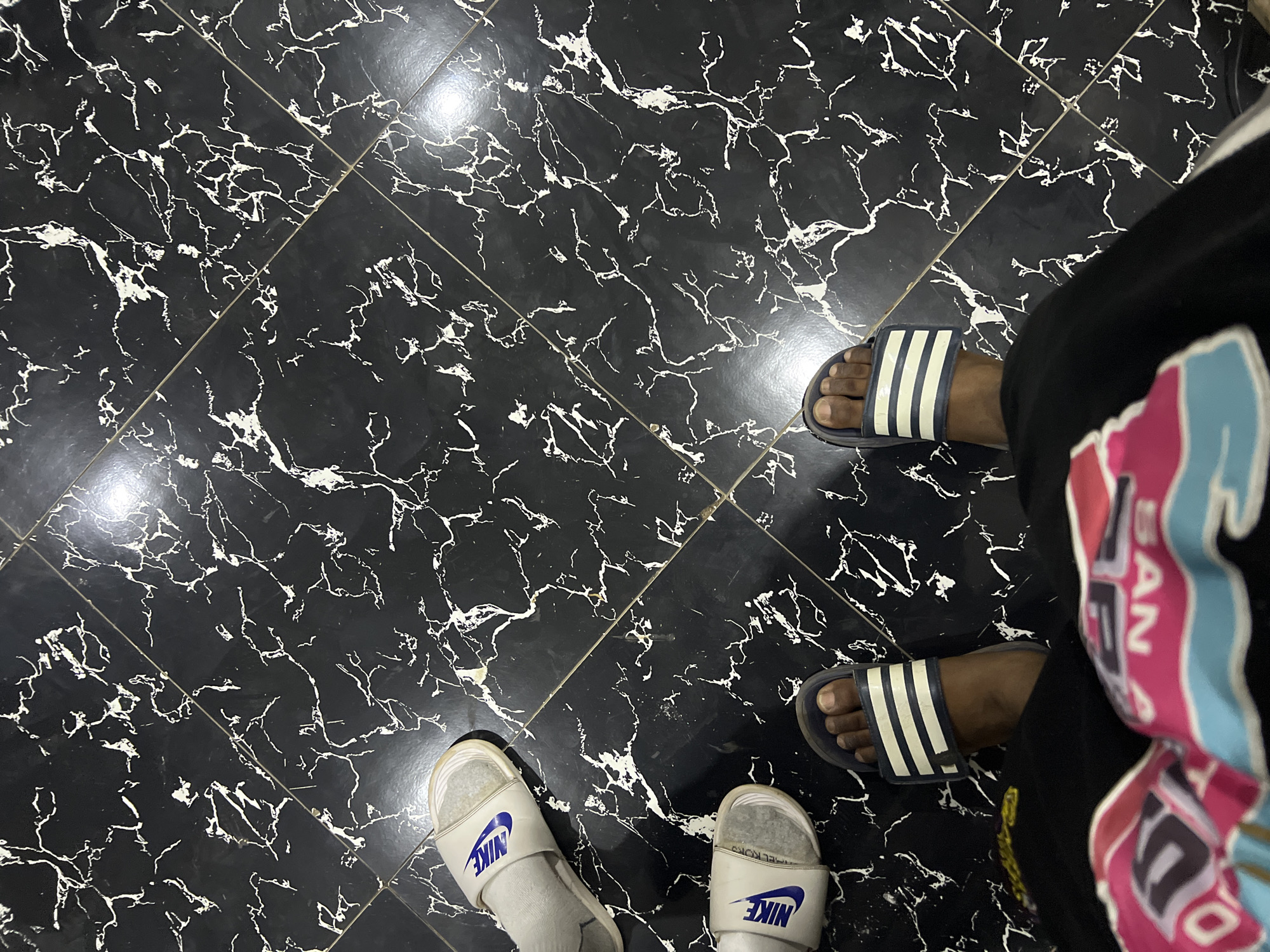 An overhead shot captures the feet of two individuals standing on a black tiled floor with a distinctive white, marble-like veining pattern. The tiles are arranged in a grid. On the left side of the frame, a person wears white Nike slides over white socks; the blue "NIKE" logo is clearly visible on the strap of both slides. On the right, another person's feet are visible in blue Adidas slides, which feature three prominent white stripes. This individual appears to be wearing bare feet in the slides, black bottoms (likely pants or shorts), and a vibrant, multi-colored garment (possibly a hoodie or t-shirt) with shades of pink, blue, and white. Partial text, including "BAN," is discernible on the colorful garment. The image depicts a casual, static moment indoors, in what is contextually identified as Abuja, Nigeria.