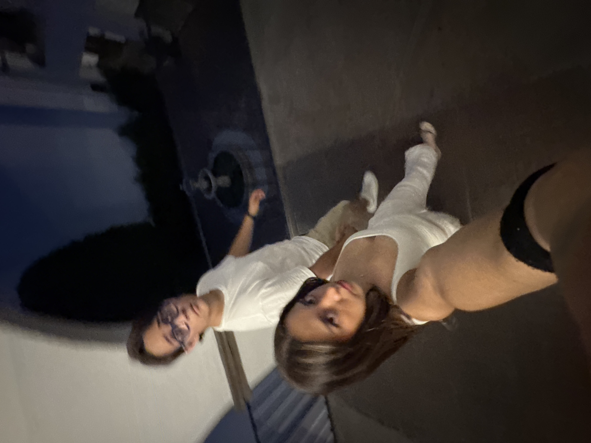 Two individuals pose for a selfie. One individual, wearing glasses and a white shirt, stands adjacent to a fountain in Cocoyoc, Mexico. A second individual, wearing a white tank top and dark bottoms, takes the photo. They are standing on pavement. The environment appears to be outdoors, possibly at night.