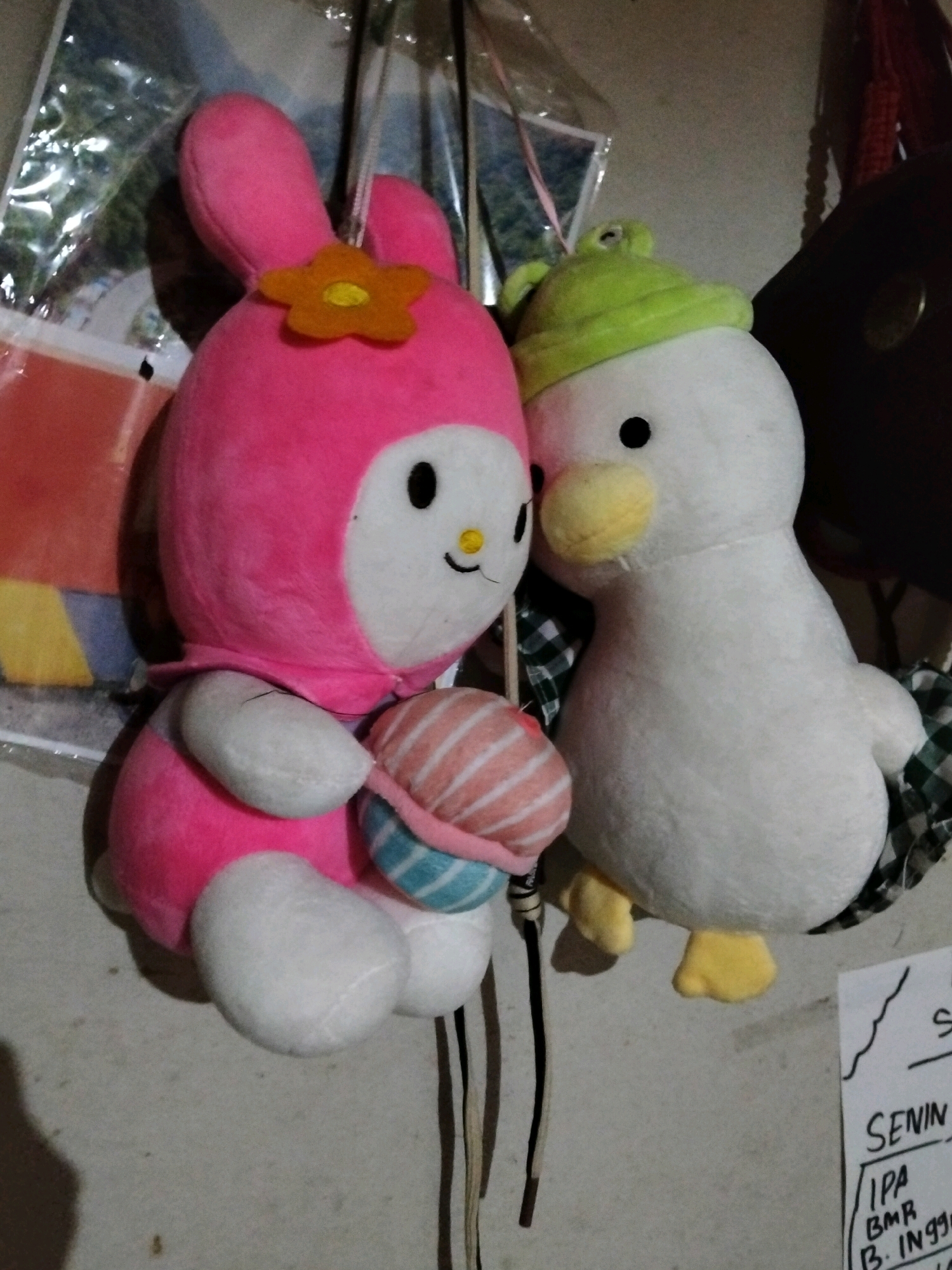 Two stuffed toys, a pink bunny-like character and a white duck, are hanging on a wall in what appears to be a domestic setting. The pink toy, resembling a character from the Hello Kitty franchise (likely My Melody), has large ears, a yellow flower accessory on its head, and is holding a striped plush object. The white duck toy is wearing a green hat and has yellow feet. The background shows a wall with some papers attached to it. The overall lighting is dim, suggesting an indoor environment. The location is identified as Teluk Pambang, Indonesia.