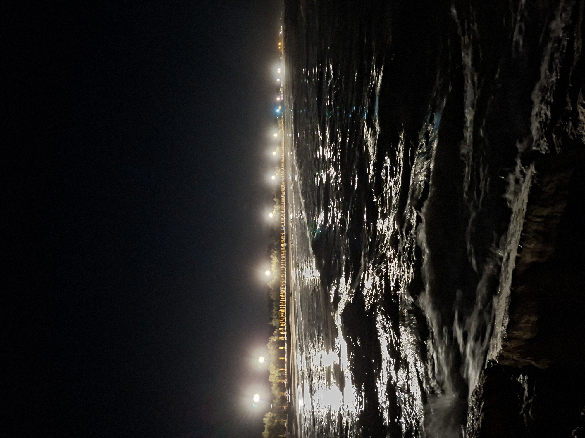 The image depicts a dark, nighttime scene of a beach in Caorle, Italy. The foreground is dominated by choppy waves crashing onto rocks, creating a frothy, illuminated effect on the water's surface due to artificial lighting. In the midground, a line of beach umbrellas and trees are visible, illuminated by a series of streetlights that stretch along the coastline. The sky above is completely dark. The lighting suggests a well-developed tourist area that is still active at night.