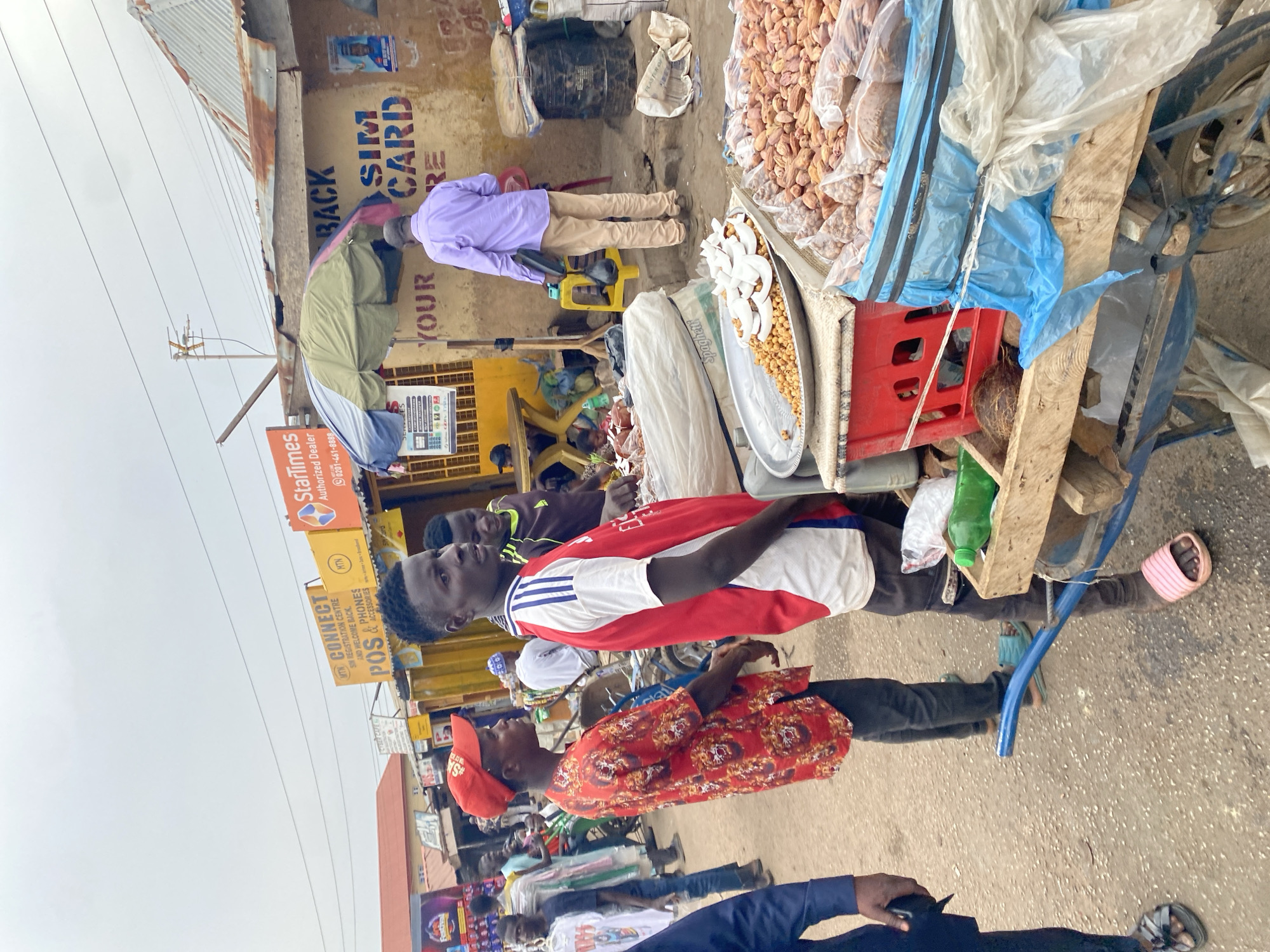 The image shows a street scene in Gura Top, Nigeria. A mobile stall displays bags of groundnuts and coconut pieces, arranged on a hand-pushed cart with a blue frame. The cart uses a red plastic crate as a platform. Nearby, an MTN "Connect" POS and phone accessory stand is visible. A man is wearing a red and white soccer jersey. Two other men stand close to the cart. A man wears a red patterned shirt and a red cap. He also wears a red "#SA" t-shirt. Another person wears a light purple shirt and light brown pants. The foreground shows a portion of the street surface.