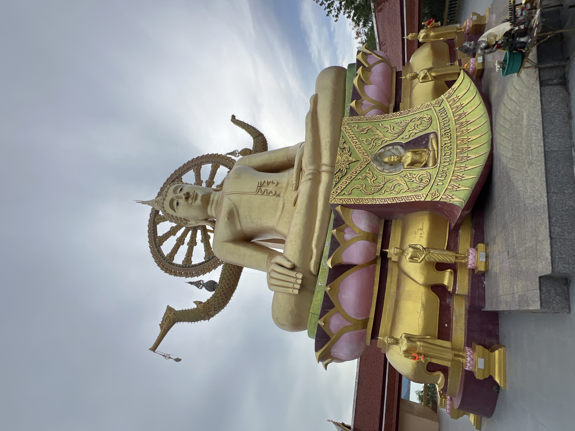The image depicts the Big Buddha of Koh Samui, located at Wat Plai Laem in Baan Plai Laem, Thailand. A massive, golden Buddha statue sits in a meditative pose on a lotus-shaped pedestal. The Buddha is adorned with traditional religious iconography, including a halo and a wheel of dharma. The statue is flanked by smaller golden statues of deities and is decorated with a vibrant, green tapestry featuring intricate golden patterns and a smaller Buddha image. The surrounding environment includes traditional Thai architecture with red tiled roofs and lush greenery. The sky above is overcast with scattered clouds. The overall scene conveys a sense of religious serenity and cultural significance.