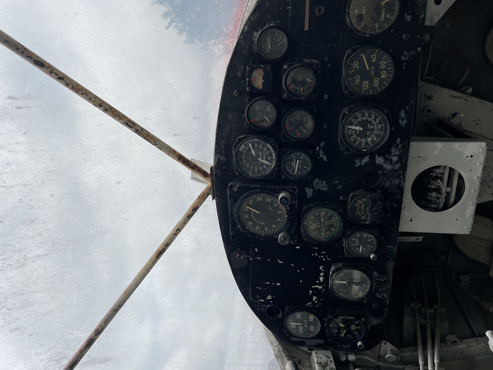 The image shows the cockpit of an old, possibly abandoned, aircraft. The dashboard is black and filled with various analog gauges and instruments, many of which appear worn and faded. These instruments include airspeed indicators, altimeters, gyroscopes, fuel quantity indicators, oil pressure gauges, and a voltmeter. Two rusty metal rods extend upwards from the dashboard towards the sky, which is cloudy and overcast. The overall environment suggests an outdoor setting, possibly in a rural or neglected area. The image was taken in the city of Na Chom Thian, Thailand.