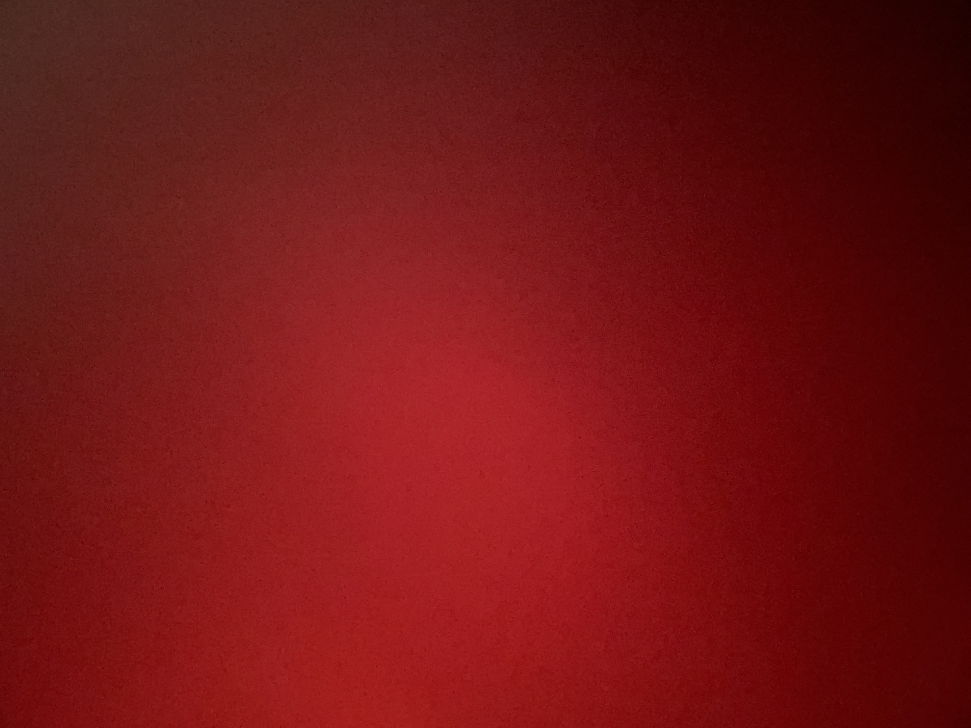 The image is a plain, textured, red background. It appears to be a solid color, possibly fabric or a painted surface, with a subtle, grainy texture visible throughout. There is a gradient of color, with the left side of the image being a darker, more muted red, and the color gradually becoming brighter and more vibrant towards the right side, culminating in a deep, almost blackish-red on the far right edge. The lighting creates a soft gradient, with no harsh shadows or bright highlights. There are no discernible subjects, objects, activities, or text in the image. The overall impression is that of a simple, abstract background.