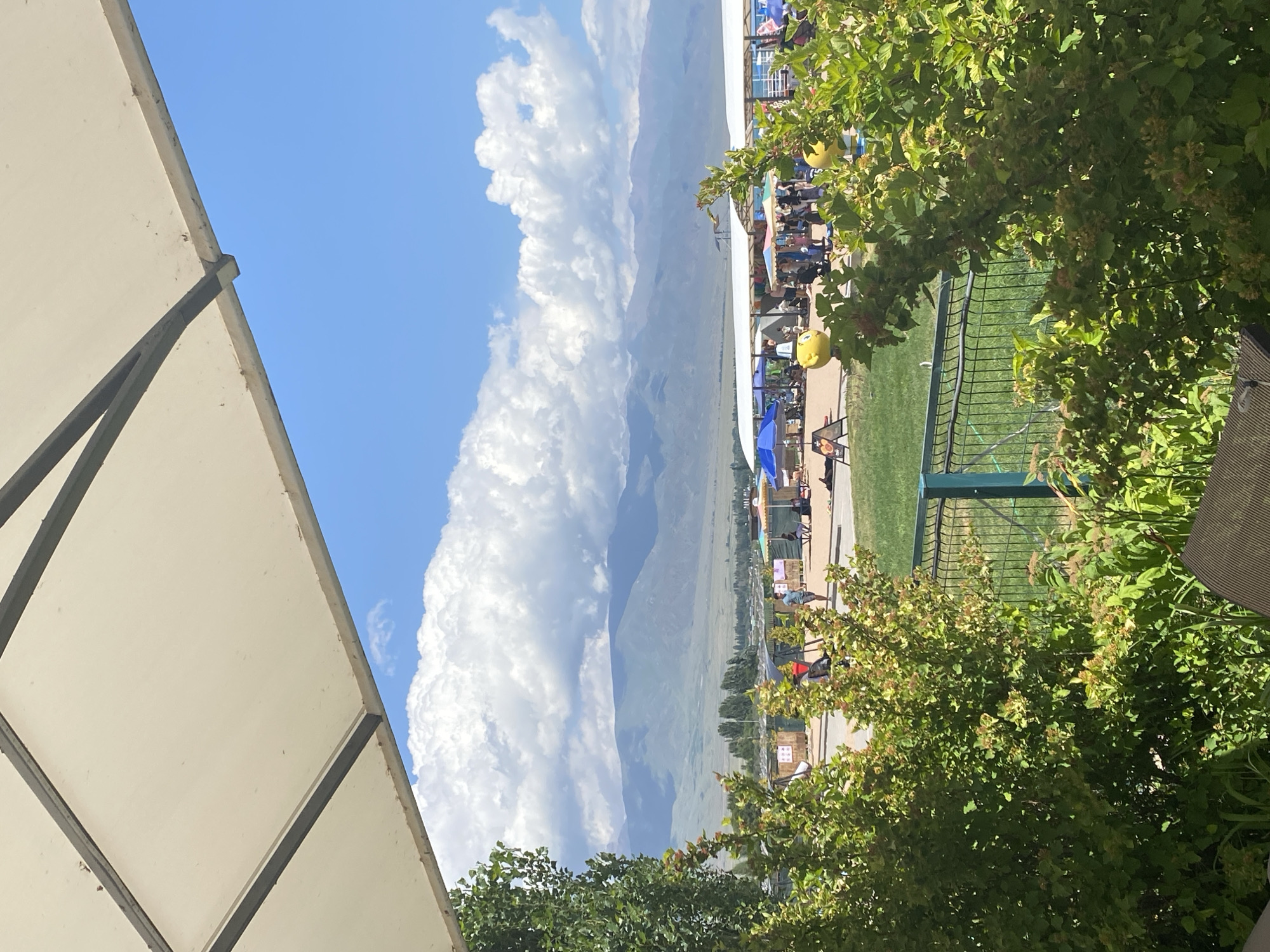 The image shows a view from under a large white awning, looking out at a sunny day with a blue sky filled with white, puffy clouds. In the background, a range of majestic, hazy mountains is visible. In the middle ground, there's a lively scene with various stalls and tents, some with blue umbrellas, suggesting an outdoor market or event. Several people are scattered around, some walking, others seated under the tents, and a few are sunbathing. There are also yellow, cartoon-like inflatable figures. A green fence separates the foreground from the middle ground. Lush green bushes with small red berries and other foliage are prominent in the immediate foreground. The scene is likely set in Koshkol', Kyrgyzstan, given the mountainous landscape.