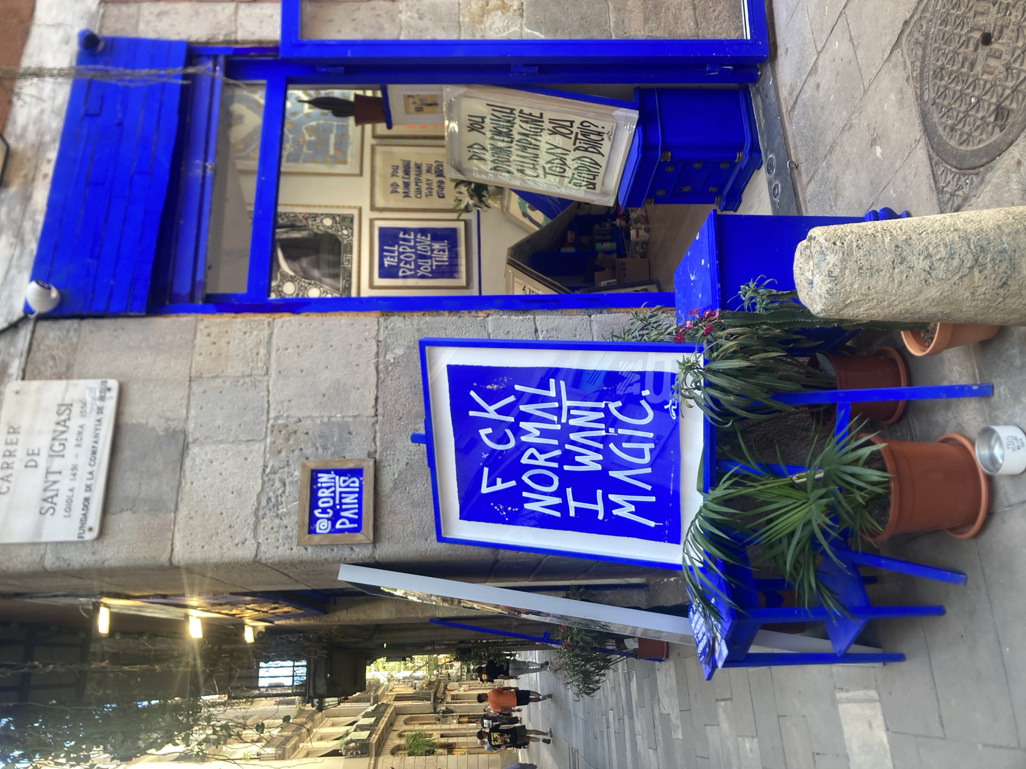 This image shows a street scene in Barcelona, Spain. The street, named "Carrer de Sant Ignasi," features a shopfront painted in a striking blue color. A sign bearing the text "FCK NORMAL I WANT MAGIC" stands outside the shop, surrounded by potted plants and a blue wooden structure. The shop window displays framed prints with phrases like "TELL PEOPLE YOU LOVE THEM" and "DID YOU DRINK ENOUGH CHAMPAGNE TODAY YOU STUPID BITCH?". A small sign on the wall identifies "@CORIN PAINTS." Several people are walking down the street in the background.
