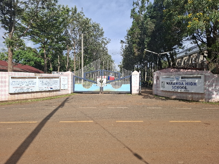 The image shows the gate entrance of the Maranda High School, located at coordinates -0.087 latitude, 34.226 longitude in Bondo, Kenya. The gate is made of metal bars, and it is painted blue and white. The school sign is a rectangular, white placard with the words "MARANDA HIGH SCHOOL" in blue lettering and the words "Ministry of Education" in smaller black lettering beneath it. The sign is mounted on a light pink wall. There are two white rectangular signs, one on either side of the gate, displaying school policies and information in black text. There is an empty paved road in front of the gate and a shadow cast by the building to the left. The road is flanked by two rows of lush trees. The sky is clear with a few puffy white clouds. There are no people visible in the image.