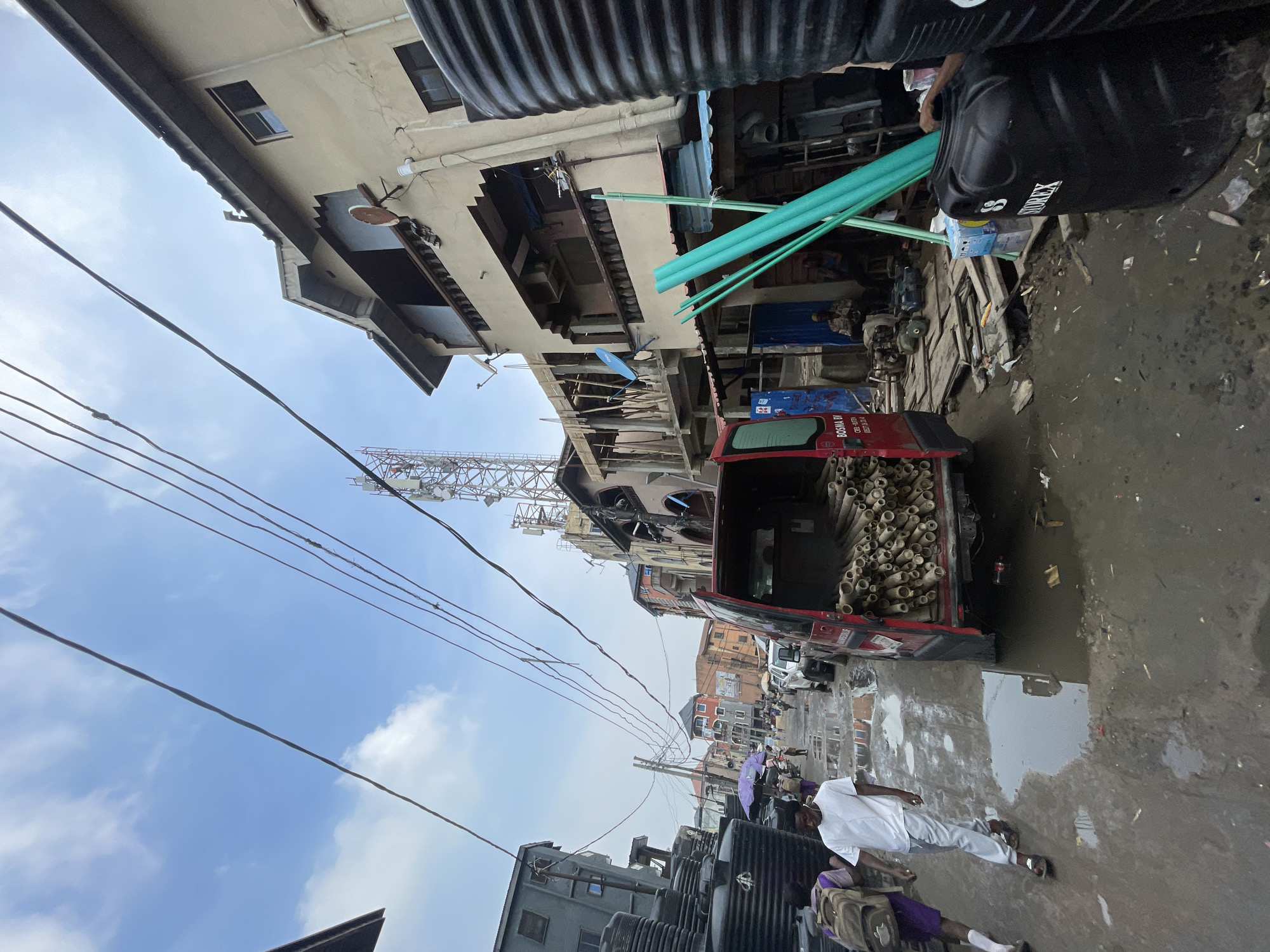 The image depicts a street in Lagos, Nigeria, filled with black water tanks and various materials. A red truck is partially visible, its bed loaded with tan-colored pipes. Several green pipes are stacked nearby. Two individuals are walking along the street, one wearing a white shirt and pants, the other in a purple top and carrying a backpack, holding a purple umbrella. The street itself appears to be partially flooded or has standing water. Buildings line the street, with a cellular tower visible in the background. Power lines crisscross above.