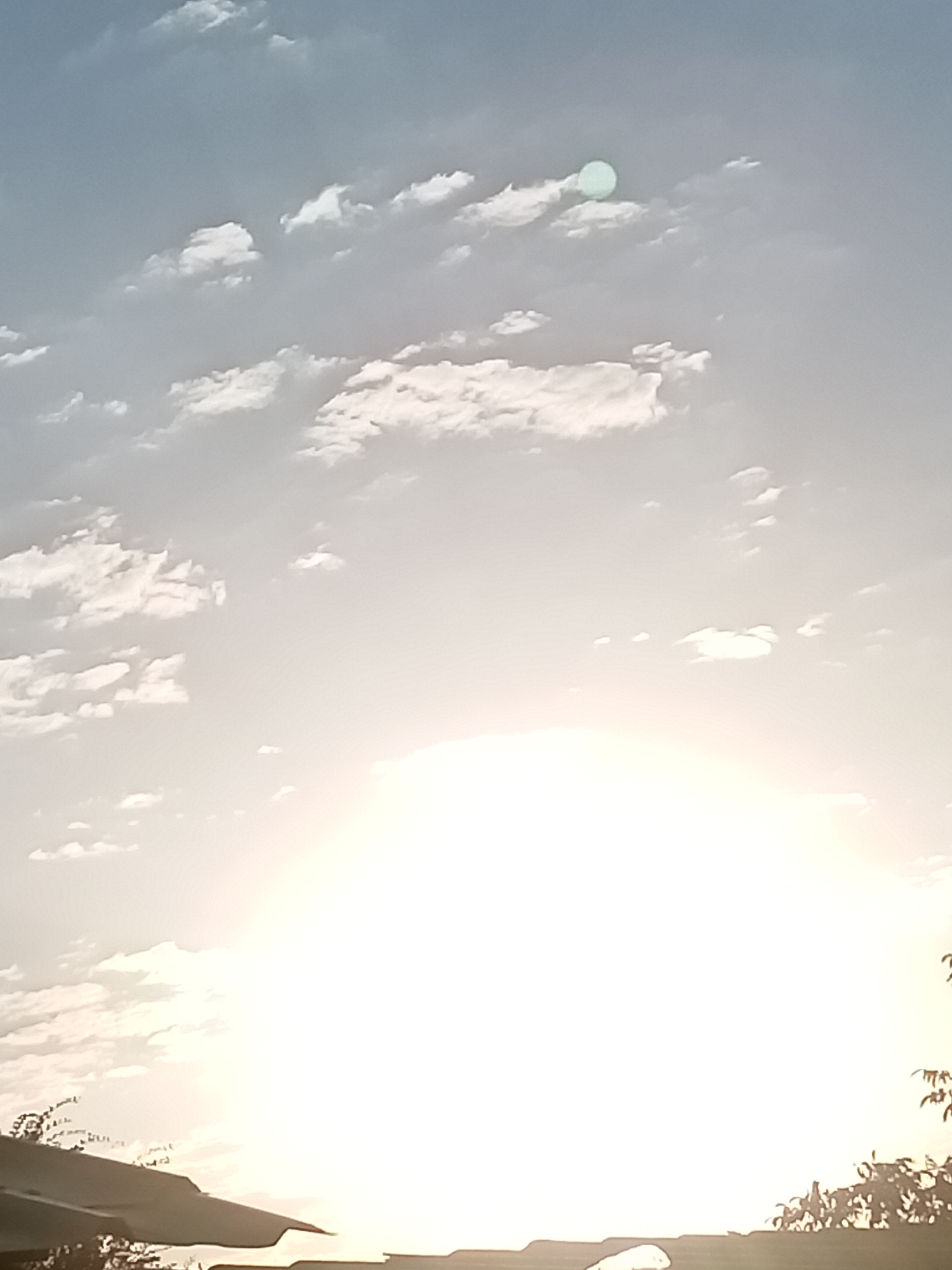 The image depicts a bright, hazy sky with scattered clouds, likely during sunrise or sunset. The sun is a dominant, overexposed bright light source at the bottom center, obscuring much of the detail in that area. Above the sun, the sky transitions from a bright white to a soft blue. A circular, pale green object, possibly a lens flare or an artifact, is visible in the upper portion of the sky, to the left of center. Silhouetted against the bright sky at the very bottom are the outlines of what appear to be rooftops and vegetation, suggesting an outdoor scene in a populated area. The overall impression is that of a sky at a time of day with intense light.