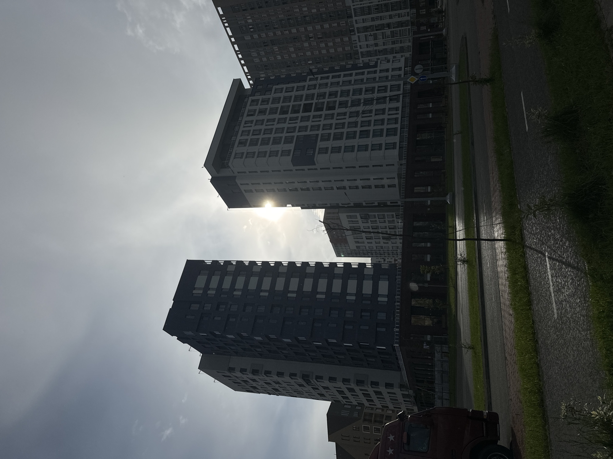 The image depicts a street scene in a city, likely a residential or mixed-use development, with several modern high-rise buildings dominating the background. The sky is overcast with the sun peeking through, creating a hazy, diffused light. In the foreground, a dark red truck is partially visible on the left side of the frame. The street is paved, with a sidewalk and a strip of green grass bordering it. The buildings are characterized by their contemporary architecture, featuring multiple windows and clean lines. One prominent building is dark blue-gray, while another is lighter in color with a more intricate facade. A third building to the far right is densely packed with windows. The overall impression is one of urban development with a focus on vertical structures.