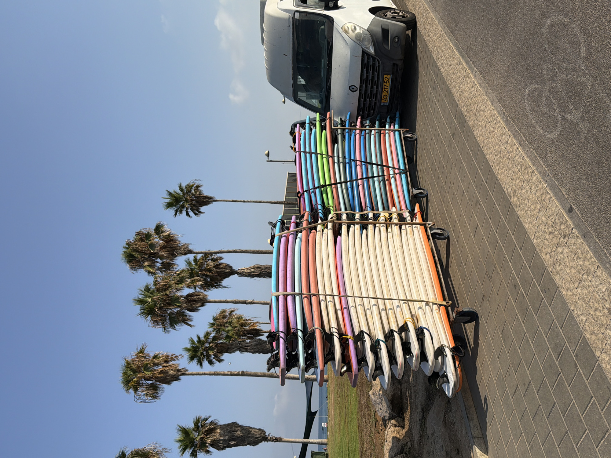 In Tel-Aviv, Israel, on a clear day, a fleet of surfboards is neatly stacked on a two-tiered cart. The surfboards are colorfully arranged with the top boards showcasing shades of blue, purple, and orange, while the lower boards are predominantly white and orange. To the right of the surfboards, a white panel van is parked. The van's license plate is visible and reads "43-217-52". The scene is set against a backdrop of palm trees and a tranquil blue sky. The foreground is paved with interlocking gray bricks, indicating a pedestrian walkway. A short distance away, a strip of green grass separates the walkway from the sandy beach and the calm sea, suggesting a location near the coastline. The overall atmosphere is one of a serene beach town ready for a day of surfing.