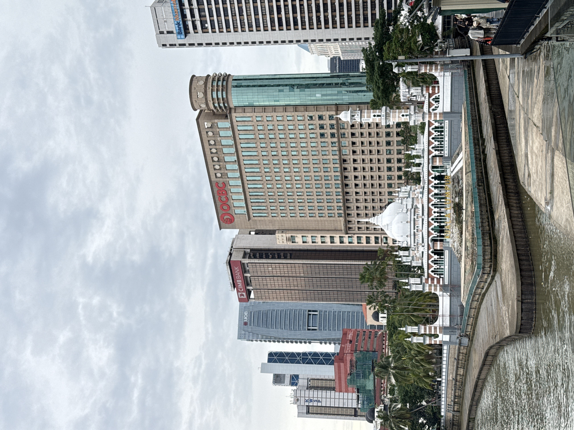 The image shows a low-angle view of the skyline of Kuala Lumpur, Malaysia, with several prominent buildings and a river in the foreground. The sky is overcast.
The buildings include:
* A tall building with the OCBC logo.
* A tall building with the CIMB Bank logo.
* A tall building with the UOB logo.
* The Sultan Abdul Samad Building.
The river is flowing, and there is a concrete embankment along its edge. Some people can be seen on the embankment and the foreground contains palm trees.