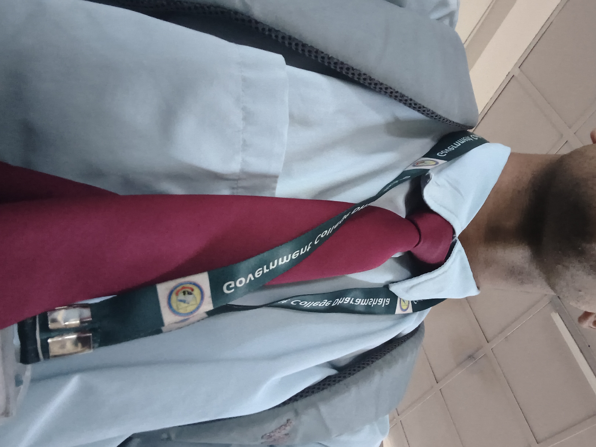A close-up, chest-up shot shows a person from the shoulders up, wearing a light blue collared shirt and a dark red tie. A dark green lanyard with text and logos is draped around their neck and over their shirt. The lanyard appears to have a school or organization emblem on it. The person is also wearing a light gray jacket or vest over their shirt, with the collar of the shirt peeking out from underneath. The background consists of a white tiled ceiling with fluorescent lighting fixtures, suggesting an indoor environment like a school hallway or office building. The lighting is somewhat dim and appears to be artificial. The person's face is only partially visible at the top of the frame, showing their chin and neck. The image is taken from a low angle, looking up towards the person.