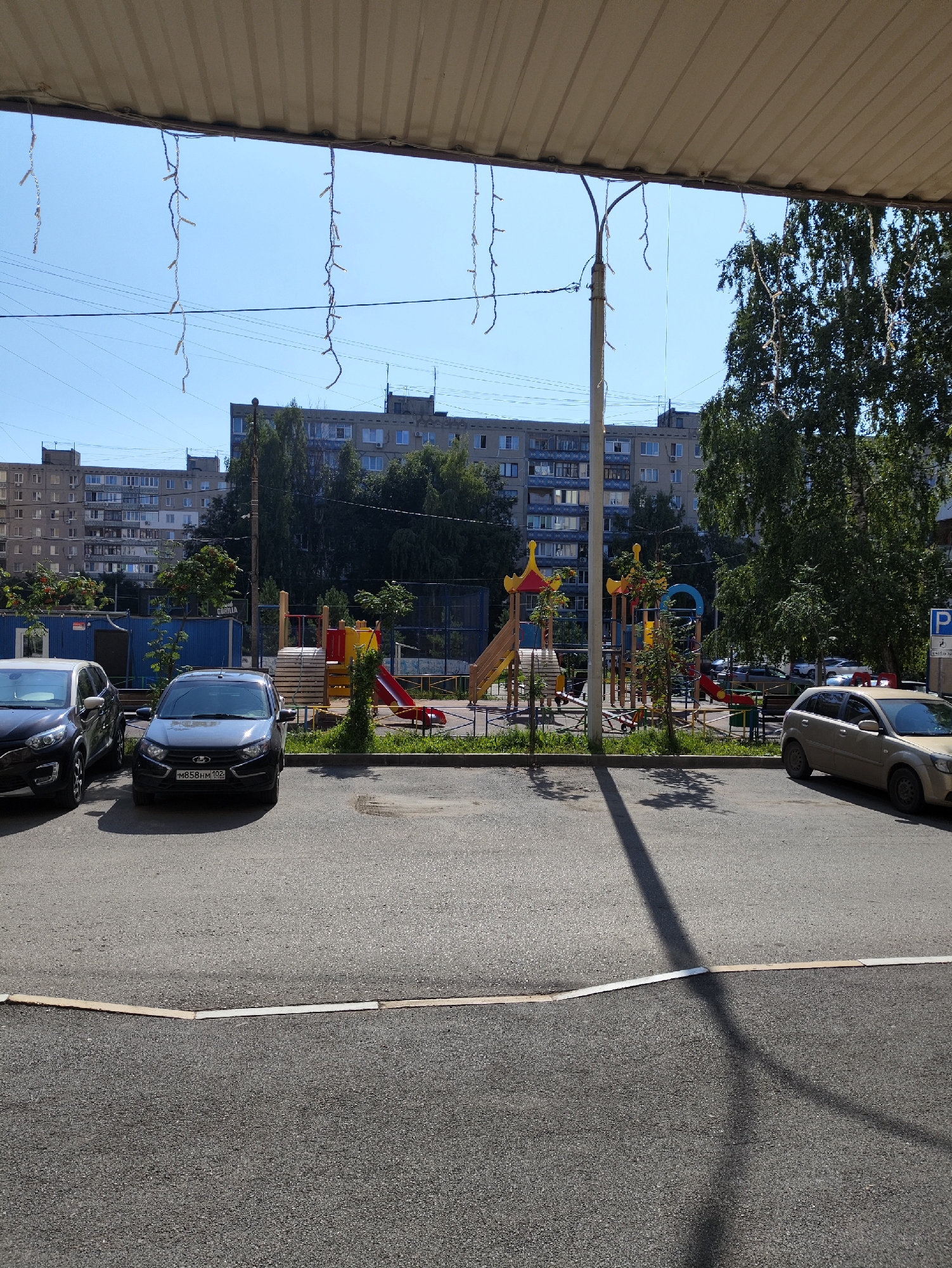 The image shows an outdoor view of a residential courtyard in Ufa, Russia. In the foreground, there is an asphalt parking lot with three cars visible. The car closest to the viewer is a dark grey sedan, followed by a black SUV. To the right, a light beige hatchback is parked. A yellow speed bump runs across the asphalt in the foreground. Beyond the parking lot, there is a playground with colorful equipment including slides and swings. Several trees with green foliage are scattered throughout the courtyard. In the background, multi-story apartment buildings are visible, characteristic of urban residential areas. The sky is clear and blue. Overhead, a corrugated metal awning extends from a building, with decorative string lights hanging from it. The scene appears to be illuminated by natural daylight.