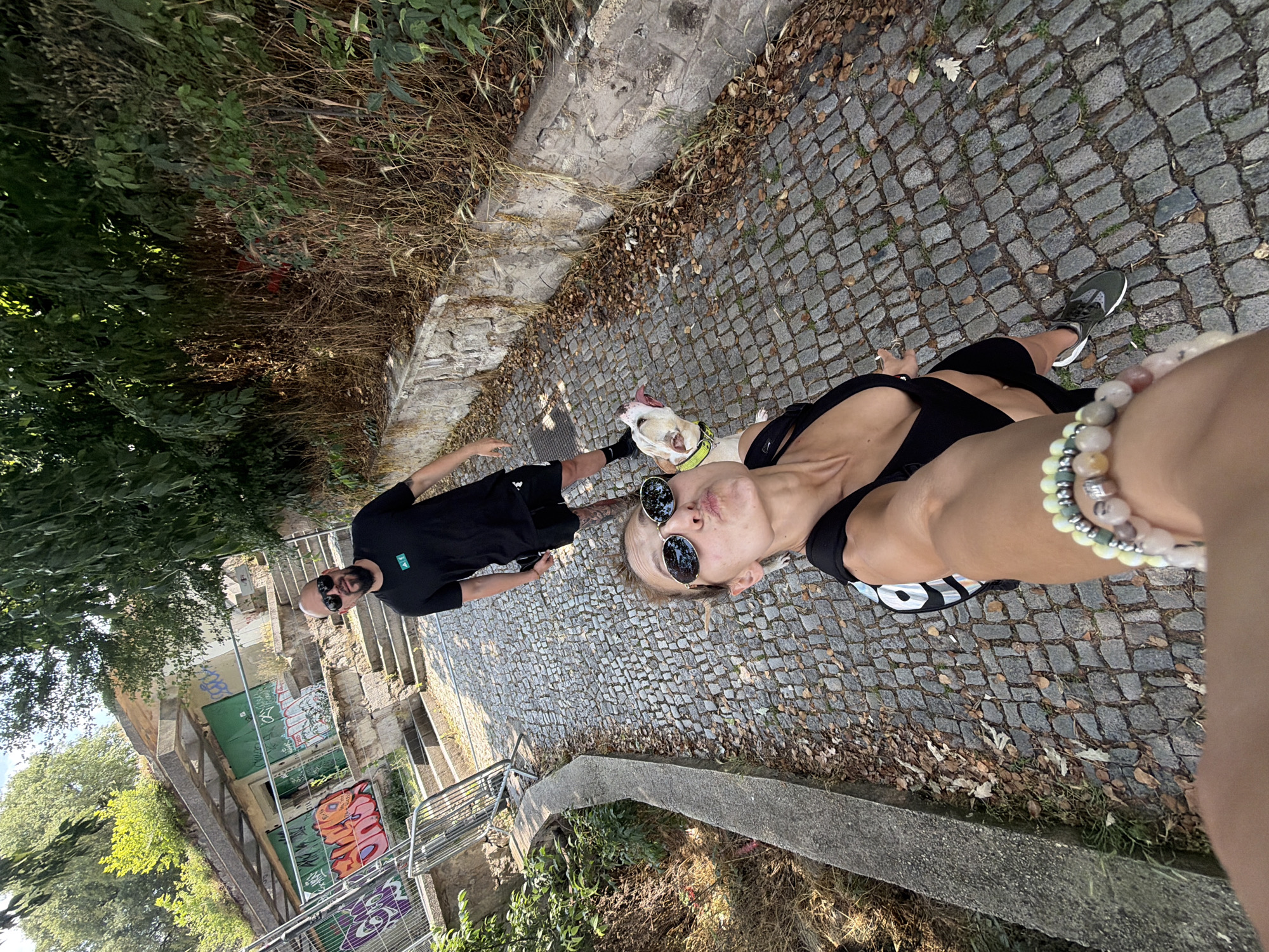 The image displays an outdoor scene on a sunlit cobblestone path in Plovdiv, Bulgaria, featuring two individuals and a dog.
In the foreground, a woman, positioned for a selfie, extends her right arm towards the camera. Her wrist is adorned with multiple beaded bracelets in shades of green, white, and pink. She wears dark oval sunglasses and a black athletic top, with her lips puckered in a "kiss" expression directed at the camera.
To her right and slightly behind, a light-colored dog, possibly a pit bull or similar breed, is visible. It wears a bright yellow-green collar and leash, and its mouth is open with its tongue extended, looking upwards towards the woman.
Further back on the cobblestone path stands a man. He has a beard and wears dark sunglasses, a black t-shirt with a small green logo on the left chest, and dark shorts, revealing tattoos on his lower right leg.
The ground is paved with irregular grey cobblestones, interspersed with scattered dry leaves. To the left, a low concrete curb borders an area of dry grass and sparse green vegetation. To the right, a low stone wall supports a dense growth of green foliage and dry, tall weeds.
In the background, behind the man, lies a dilapidated concrete and stone structure. This building features multiple concrete steps leading upwards, and its walls are extensively covered in multi-colored graffiti, including sections painted green. Metal security barriers or fences are erected in front of the lower portion of the structure. The roof and base of the building exhibit significant overgrowth of green vegetation. The sky is visible above the structure, indicating bright daylight conditions.
