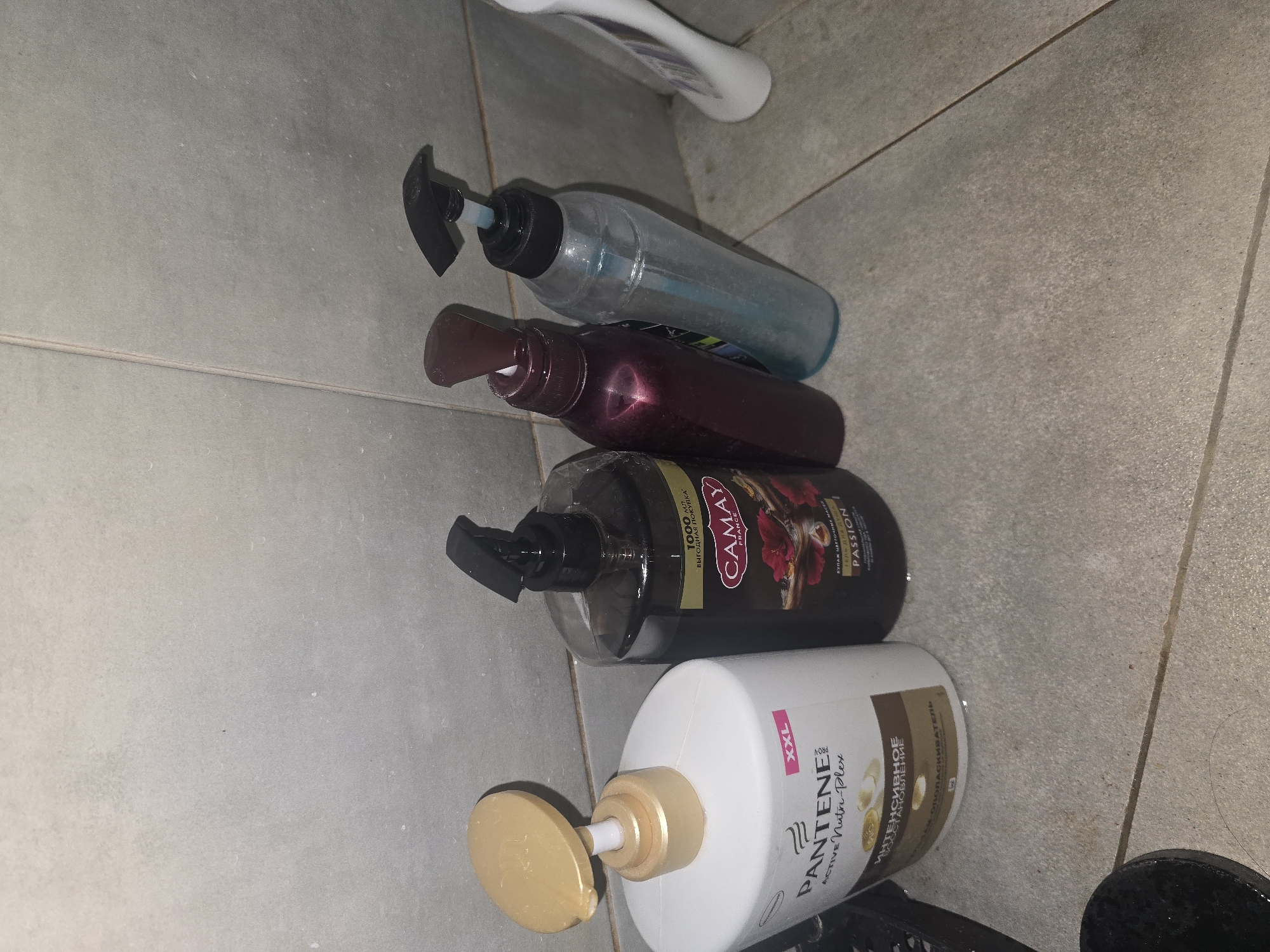 Four bottles of shampoo and conditioner are standing on a tiled floor in a bathroom. The bottles are of varying sizes and colors. From left to right, the first bottle is a large white Pantene bottle with a gold pump dispenser. The second bottle is a dark colored bottle with a black pump dispenser and a label that reads "Camay France". The third bottle is a translucent red bottle with a dark colored pump dispenser. The fourth bottle is a translucent blue bottle with a black pump dispenser. The tiles on the floor are light gray and appear to be wet. The background is a tiled wall, also light gray. The scene is consistent with a bathroom setting in Ulan-Ude, Russia.