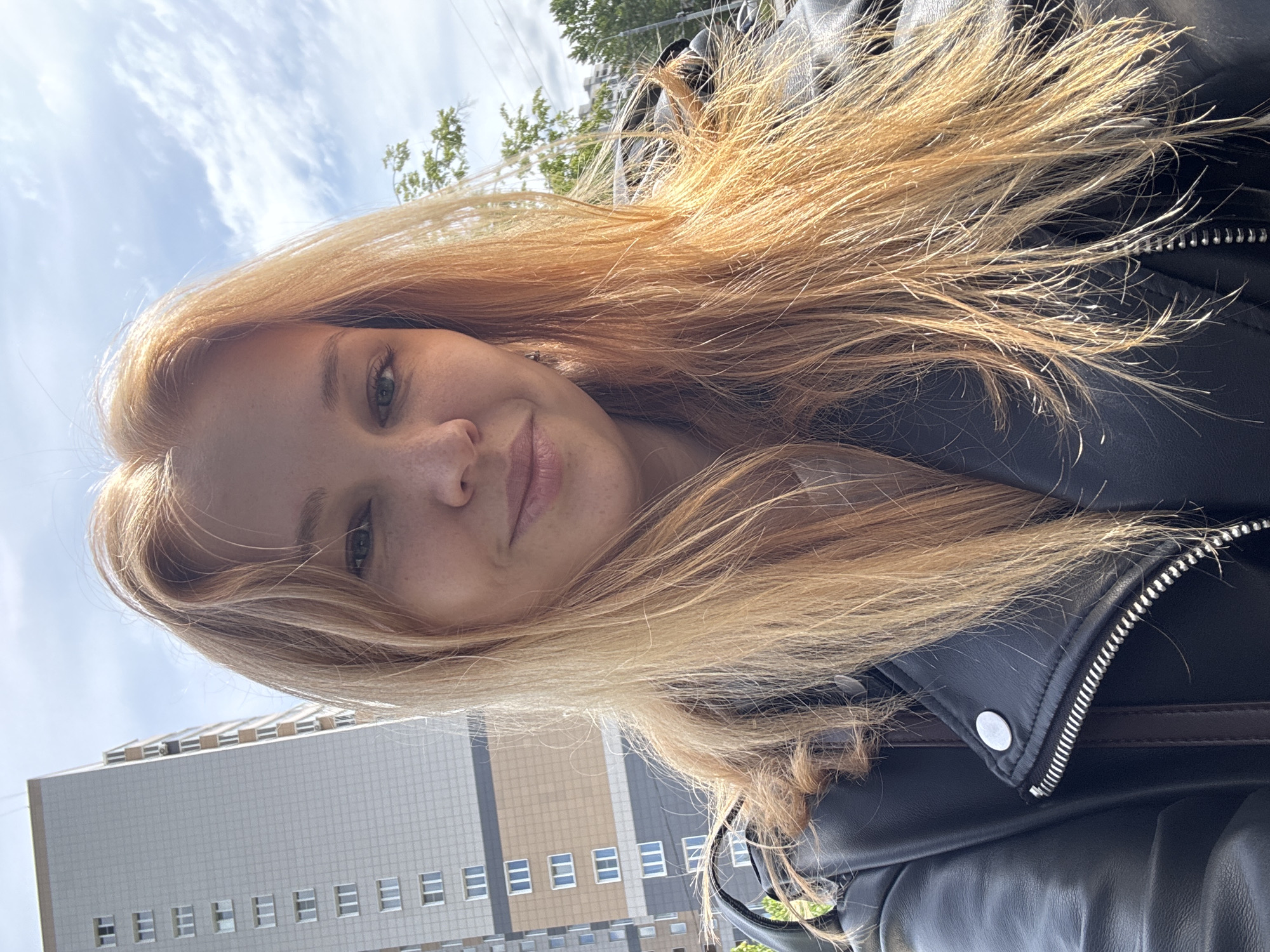 The image is a selfie of a woman with long, strawberry blonde hair. She has fair skin and some freckles on her nose and cheeks. Her eyes are light-colored. She is wearing a black leather jacket with a zipper.
In the background, there is a multi-story building with a gray and beige facade, and a partly cloudy sky. There are some trees visible as well. Based on the building style, the image is likely in the Rusanovo district in Russia.