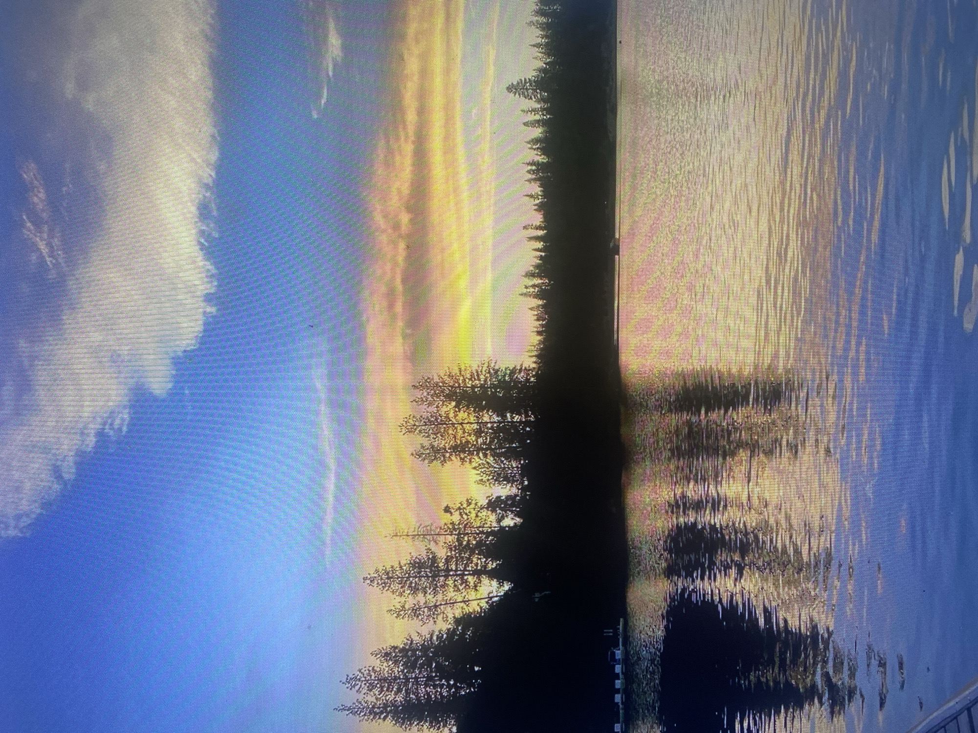 A digital photograph captures a sunset over a serene body of water, with a silhouetted treeline along the distant shore. The sky is a vibrant gradient of blues, yellows, and oranges, with wispy clouds catching the last rays of sunlight. The water reflects the colorful sky and the dark forms of the trees, creating a mirror image on its rippling surface. On the left side of the frame, a small dock with several white structures is partially visible, jutting out into the water. The overall scene evokes a sense of tranquility and natural beauty. This image does not depict the city of Samara, Russia.