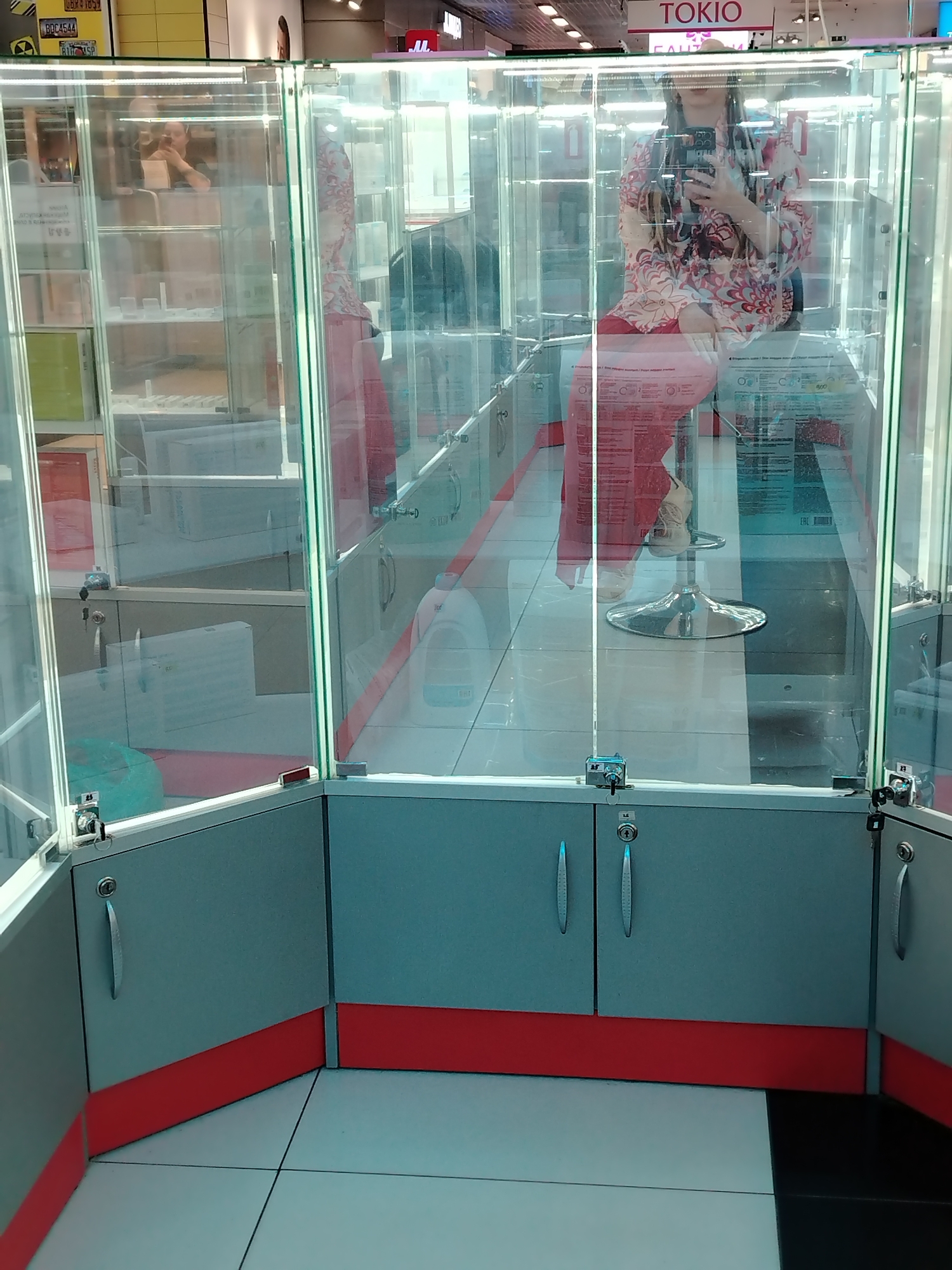 The image depicts a person seated inside a glass display case. The individual, who appears to be female, is holding a smartphone and looking at it, possibly taking a selfie. She is wearing a floral-patterned top and red pants. The display case is part of a commercial setting, likely a store, with the word "TOKIO" visible above her. The case contains several shelves with objects displayed on them. The base of the display case is grey with a red stripe along the bottom. The flooring is tiled in a black and white pattern. The specific location is identified in the metadata as being within the city of Izhevsk, Russia.
