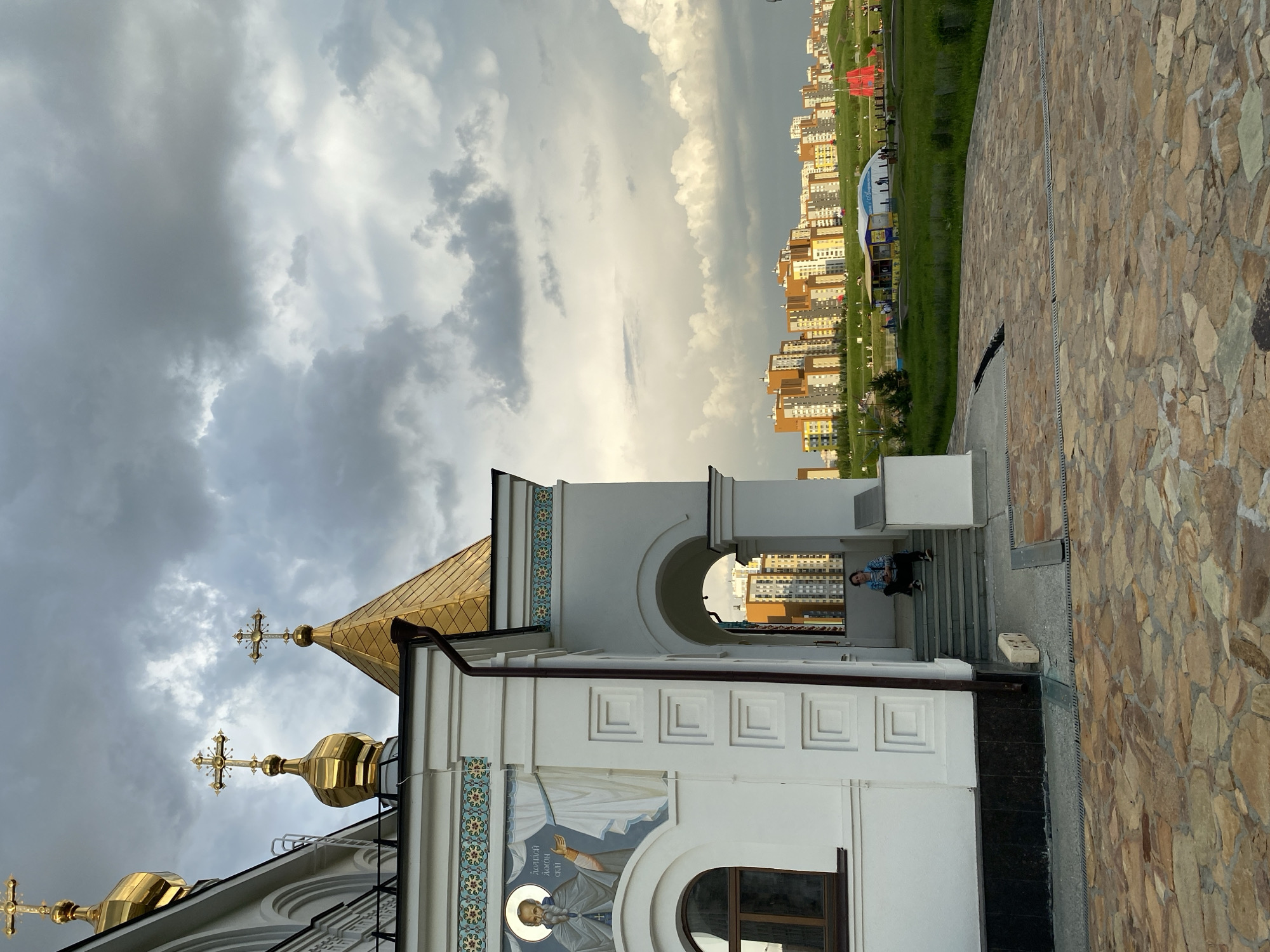 The image depicts the Church of the Archangel Michael in Yekaterinburg, Russia, set against a dramatic cloudy sky and a backdrop of modern urban development. The church, painted white with gold domes and crosses, features detailed religious artwork above its entrance. A young woman is seated on the steps of the church, looking towards the camera. Beyond the church, a park-like area with scattered people and recreational structures, including a red tent-like object, is visible, leading to a dense cityscape of multi-story buildings in various colors. The foreground is paved with irregular stone slabs.