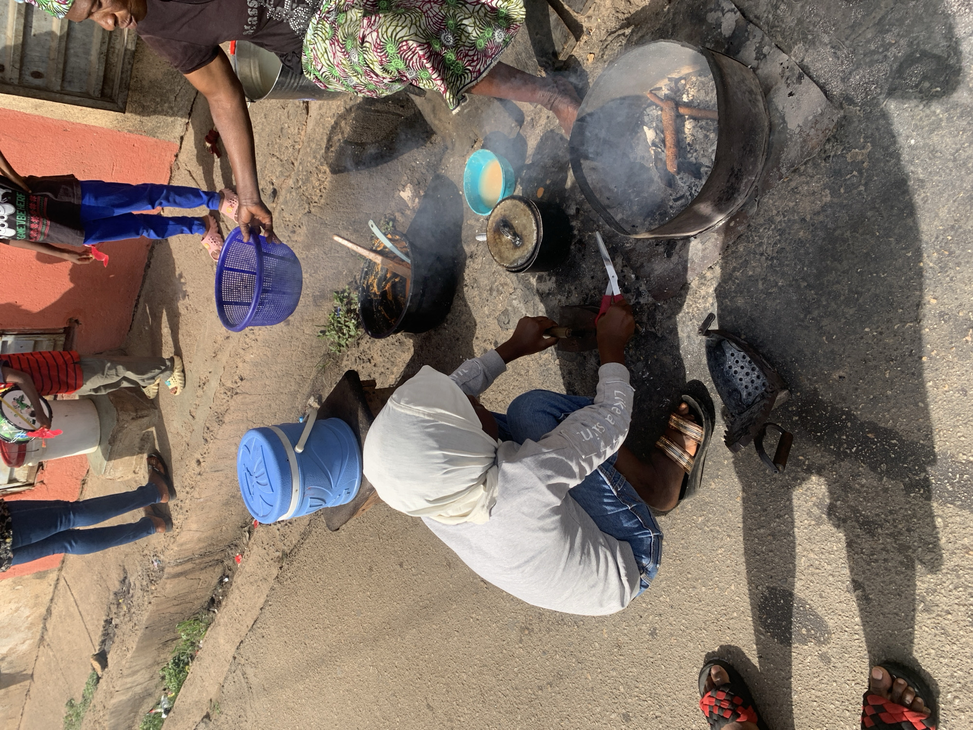 This high-angle, overhead shot captures an active outdoor scene on a concrete surface in Jos, Nigeria, likely involving food preparation. Bright sunlight casts strong, elongated shadows, suggesting a clear day in the morning or late afternoon.
Several individuals are gathered. In the upper left, a woman in a dark top and a green and white patterned skirt holds a blue plastic basket. A child in blue pants, a dark t-shirt with a white graphic (possibly Mickey Mouse ears), and pink sandals stands near a red-painted building wall. To the left, a person in a red striped shirt and grey pants is partially visible, seated.
In the center-right, a person wearing a white head covering, a grey long-sleeve shirt, and blue jeans is seated on the ground. The text "LIKE A SIN" is visible on the sleeve of their shirt. This individual is actively working with a pair of scissors and another tool, tending to an outdoor cooking setup.
The cooking area features two large, dark metal containers. One on the right is an active fire pit containing burning wood or charcoal and emitting smoke. To its left, another pot appears to hold food, with a utensil inserted. A smaller lidded pot and a light blue bowl containing a yellowish liquid are also on the ground. A blue plastic cooler with a white handle is positioned further left. Various sandals and shoes are visible, including red and black patterned sandals in the foreground, likely belonging to the photographer. The scene portrays a communal, everyday outdoor activity.