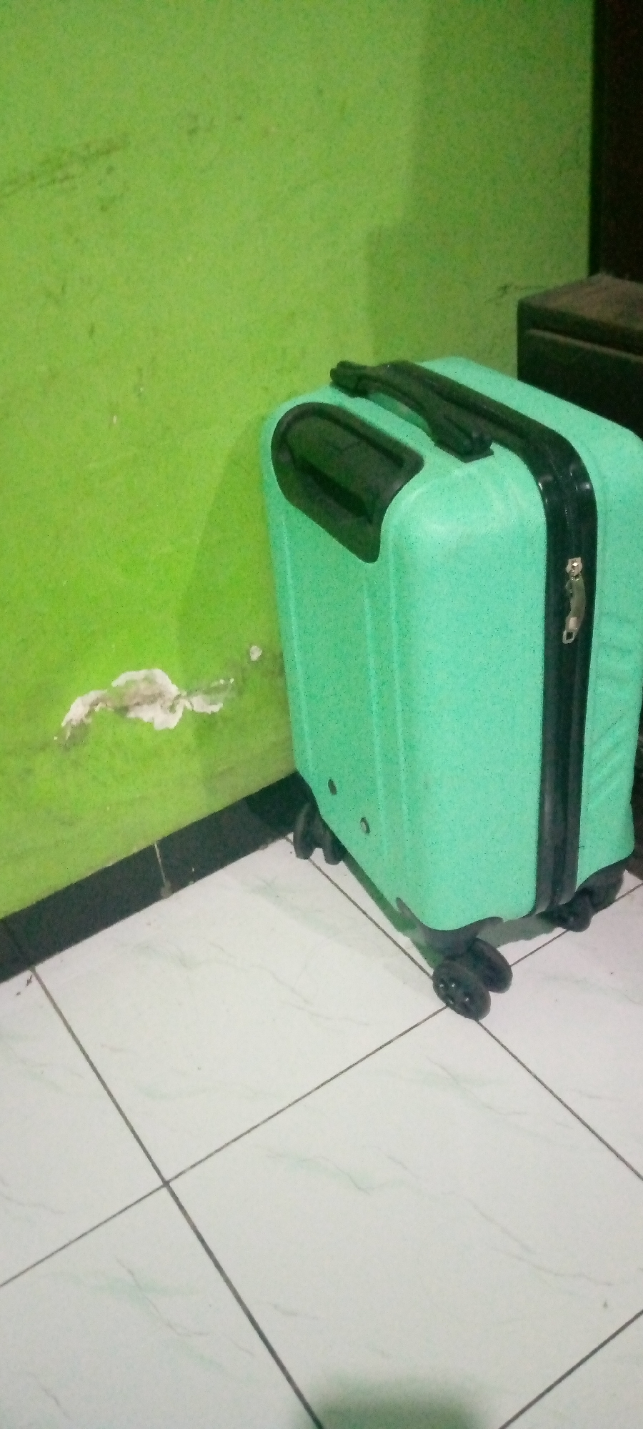 A bright green suitcase with black trim and wheels sits on a tiled floor next to a green wall. The wall has a patch of peeling paint near the floor. The scene appears to be indoors, possibly in a room within a building. The lighting suggests it may be daytime, but the room itself is not brightly lit. The suitcase is upright and stationary. There are no people or other objects clearly visible in the immediate vicinity of the suitcase. The image is taken from a medium distance, showing the suitcase and a portion of the surrounding environment.
