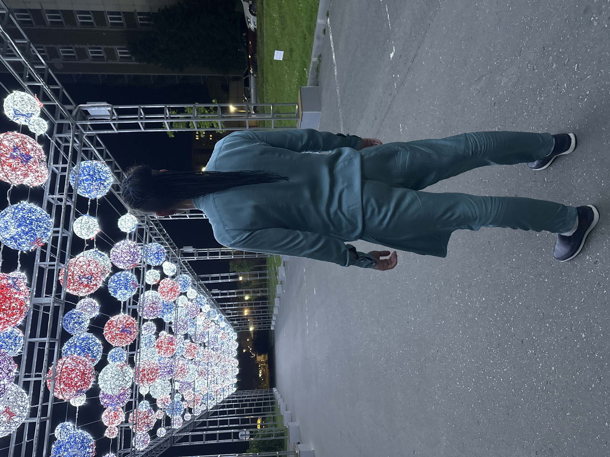 A person stands in the middle of an asphalt pathway, facing away from the camera. The pathway is lined with large, spherical lights in red, white, and blue, creating an archway. The person is wearing a teal-colored suit, with a long braid of dark hair extending down their back. In the background, there is a building with many windows, trees, and parked cars. The scene appears to be an outdoor public space at night, possibly a park or a city street decorated for an event. The image is set in Saratov, Russia, as indicated by the prompt.