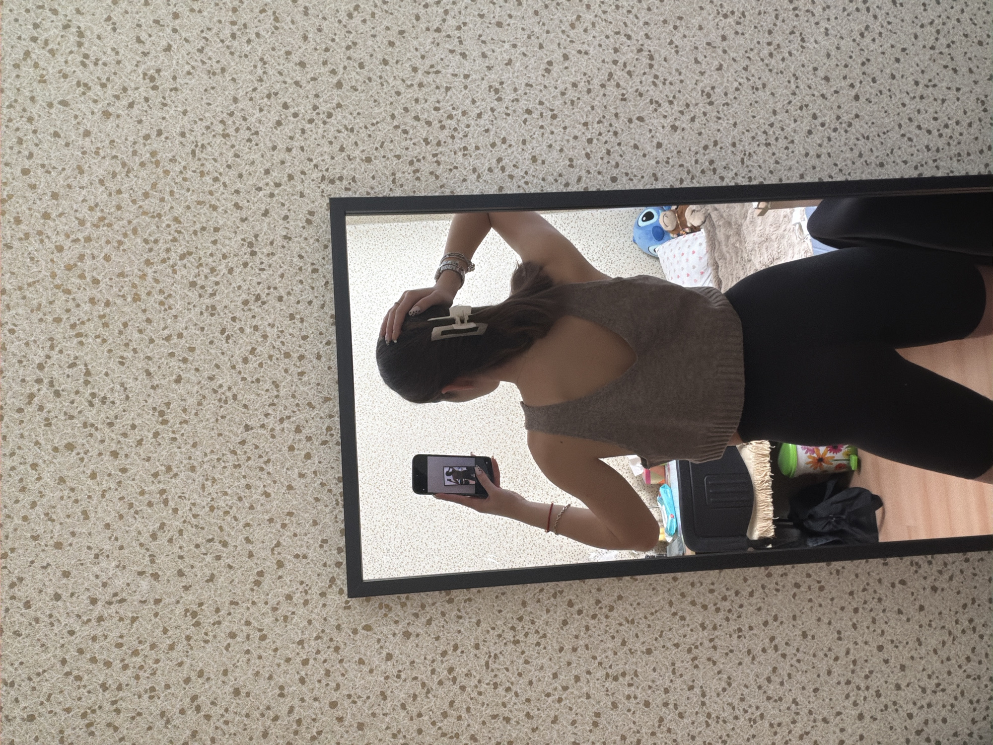 A young woman is taking a mirror selfie in an interior space. The woman is wearing a light brown tank top, black leggings, and a white hair clip. She is holding a smartphone in her right hand to capture the image. The reflection shows a cluttered room with a bed, pillows, a stuffed animal, and various other objects. The wall behind the mirror has a textured beige and white pattern. There is no information to indicate this picture was taken in Sofia, Bulgaria.
