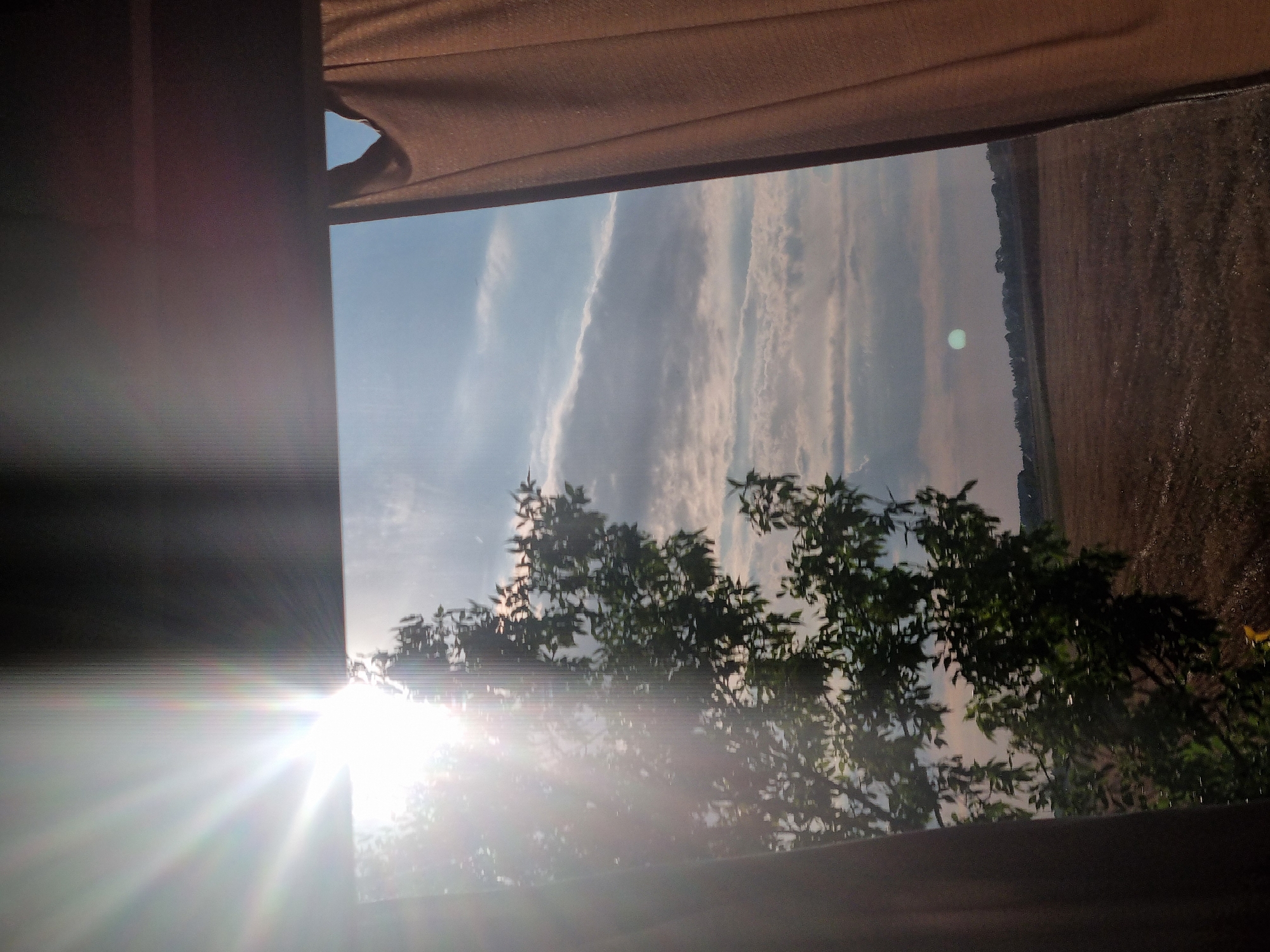 This is a photograph taken from inside a building, looking out a window. The view is of a rural landscape under a cloudy sky with bright sunlight. The sun is positioned in the upper left quadrant of the image, causing lens flare and bright rays that streak across the scene. A tree with green foliage is visible in the left middle ground, silhouetted against the bright sky and partially obscured by the sun's glare. The middle ground also shows a field with harvested crops, appearing as a textured brown expanse. A line of trees forms a distant treeline on the horizon, with a few buildings visible among them. A faint, possibly artificial, green light source is present in the sky in the right middle ground. Two curtains, one on the left and one on the right, frame the window view, with the right curtain being more prominent and a light brown color. The image appears to be taken during daylight hours, likely late afternoon or early evening given the angle of the sun. There are no people visible in the image. The image does not contain any elements that would allow for specific identification of its location within Berettyóújfalu, Hungary.
