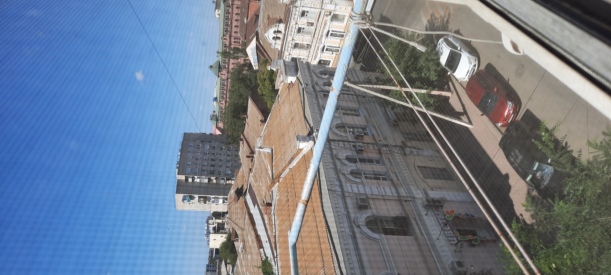 The image shows a view from an upper floor, looking down at a street and rooftops in Astrakhan, Russia. In the foreground, there is a blue pipe running horizontally across the image. Below this pipe are several buildings with rusted, corrugated metal roofs. One building in the middle ground has light-colored walls with arched windows and air conditioning units. A sign with Cyrillic lettering, "ЦВЕТЫ" (flowers), is visible on this building, indicating a flower shop.
Further back, a taller, grey apartment building with many windows stands out. To the right of this building, a pink building with green roof details and arched windows is visible. The sky is clear and blue, with a few faint clouds. On the street below, there are three cars: a black car partially obscured by greenery, a red hatchback, and a white hatchback. The scene appears to be a typical urban streetscape during the daytime.