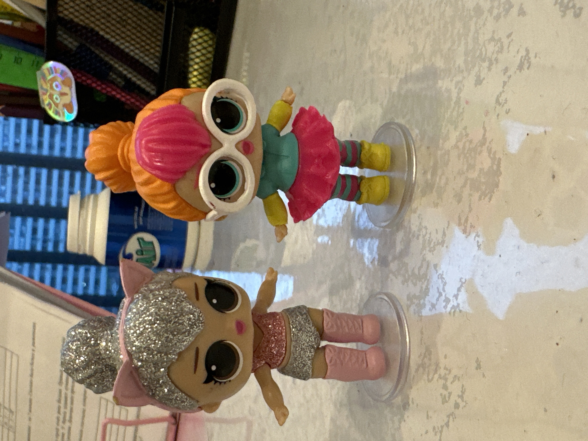 Two LOL Surprise dolls are displayed on a desk. The doll on the left has silver glitter hair with pink cat ears, a pink glitter top, a silver glitter skirt, and pink boots. The doll on the right has orange hair with a pink pompadour, large white-rimmed glasses, a teal and yellow shirt, a pink tutu skirt, and striped leggings with yellow boots. Both dolls are standing on clear circular bases. In the background, a blue and white bottle with a red and blue label is visible, along with a black mesh organizer holding some items, including a colorful sticker. The surface they are on appears to be a speckled desk or table. The image does not contain any information to suggest a specific location in Minsk, Belarus.