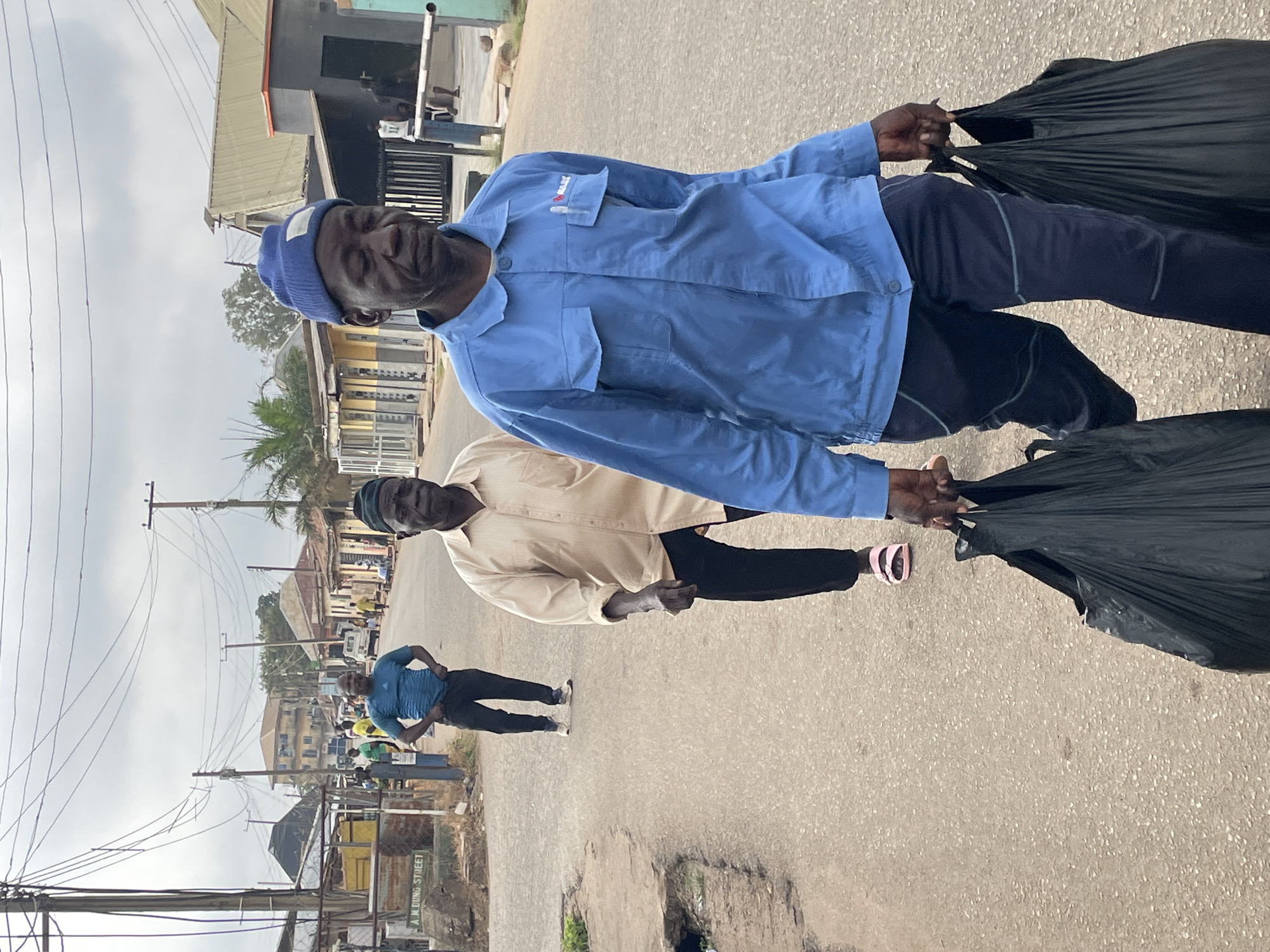 Two men carry large black trash bags down a paved street in Jos, Nigeria. The man closest to the camera wears a blue work shirt and beanie. The other man wears a tan shirt. Both men wear dark pants. A third man stands with his hands on his hips further down the street. Buildings and power lines are visible in the background. A sign on the left side of the road reads "A.M. Dung Street".