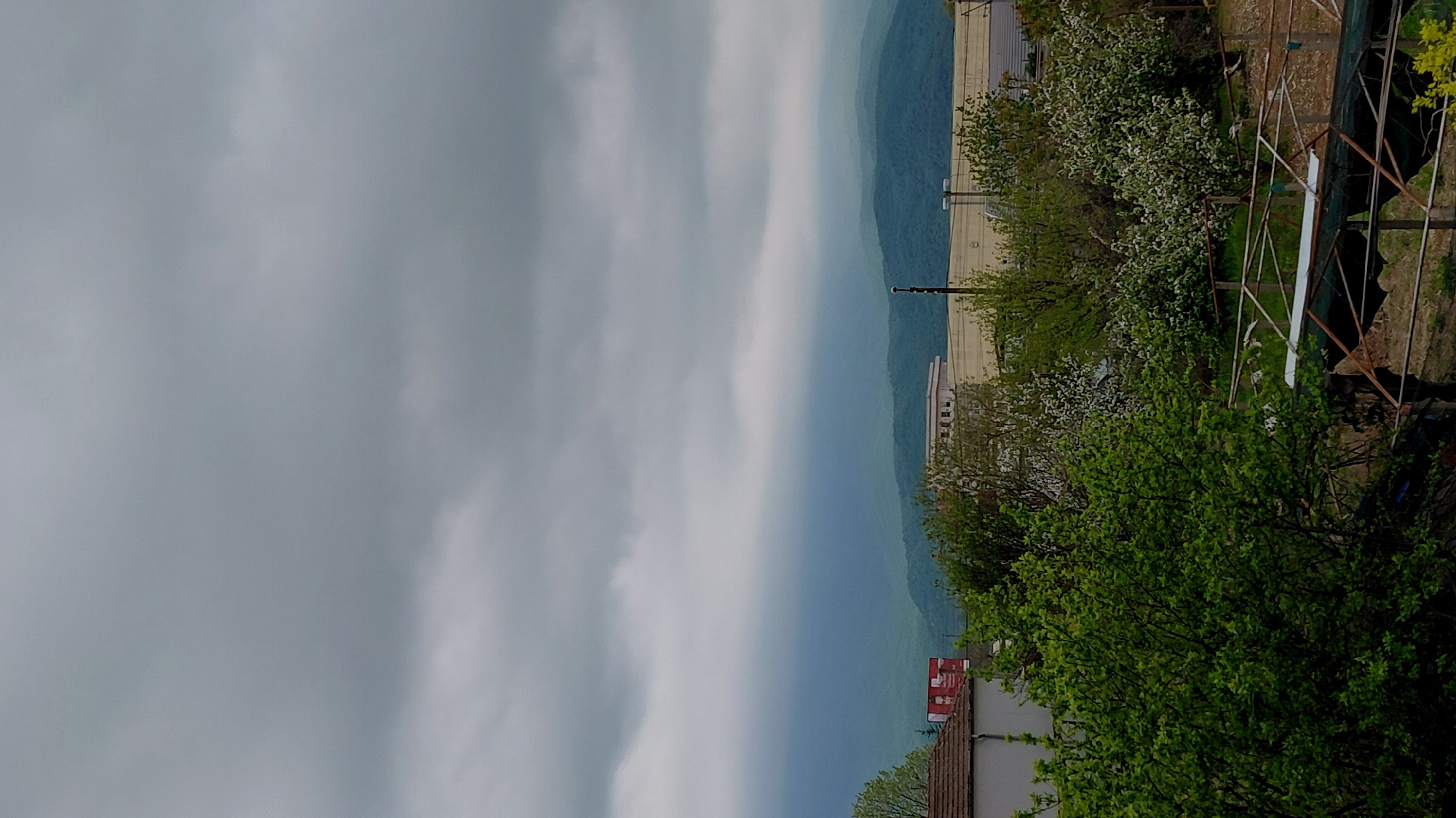 The image captures a landscape with a foreground featuring green foliage, a roof with red accents, and metal structures. Beyond this, a building stands amidst blooming trees with white flowers. In the distance, a muted mountain range is visible under an overcast sky dominated by gray clouds.