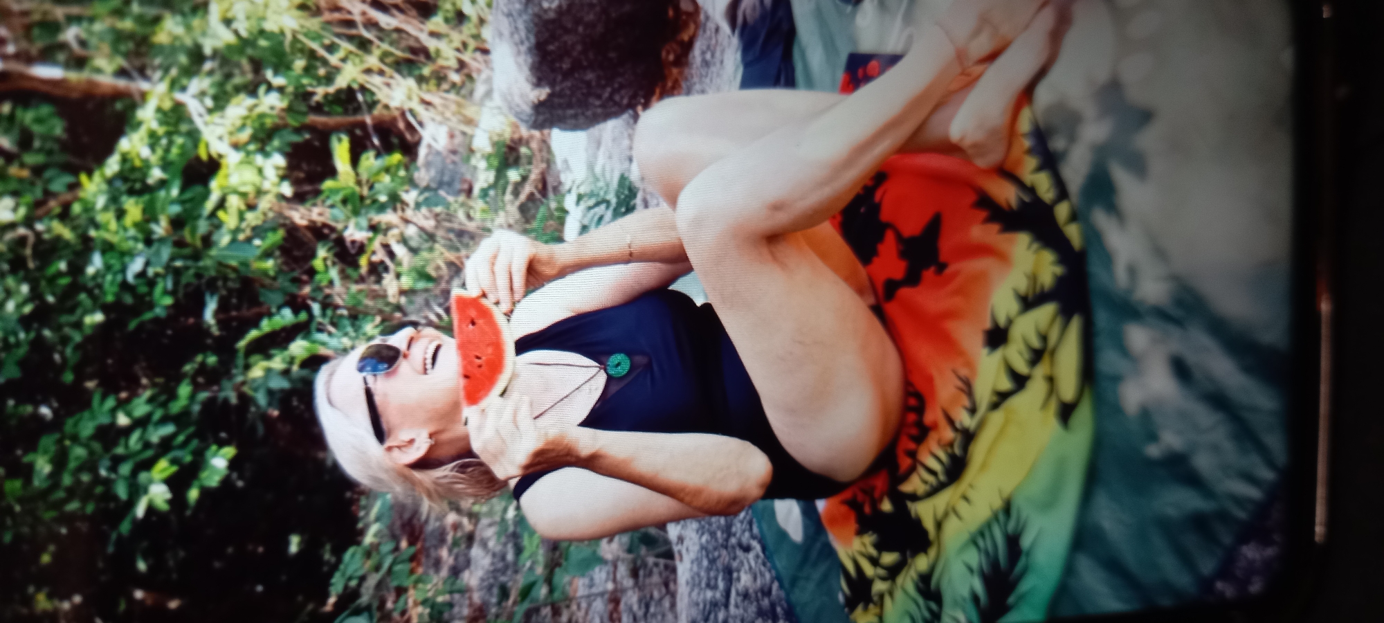 An outdoor, slightly desaturated image shows a fair-skinned woman with blonde hair and sunglasses, laughing or smiling broadly as she holds a slice of watermelon to her mouth. She is reclined or lounging, with her legs bent and a colorful, patterned fabric (likely a blanket or sarong) draped over them and underneath her. The fabric features vibrant orange, yellow, and green sections with dark silhouettes resembling palm trees or tropical foliage.
The woman is wearing a dark blue or black top with a small, round green embellishment or button visible near the neckline. She has a thin bracelet on her left wrist. The background consists of lush green foliage and what appears to be a large rock or tree trunk, suggesting a natural, possibly tropical environment. The lighting indicates it is daytime, likely sunny and warm. Her open mouth and expression convey a sense of joy and relaxation. There is no visible text in the image. The setting appears to be a tranquil spot in Baan Chaloklum, Thailand.